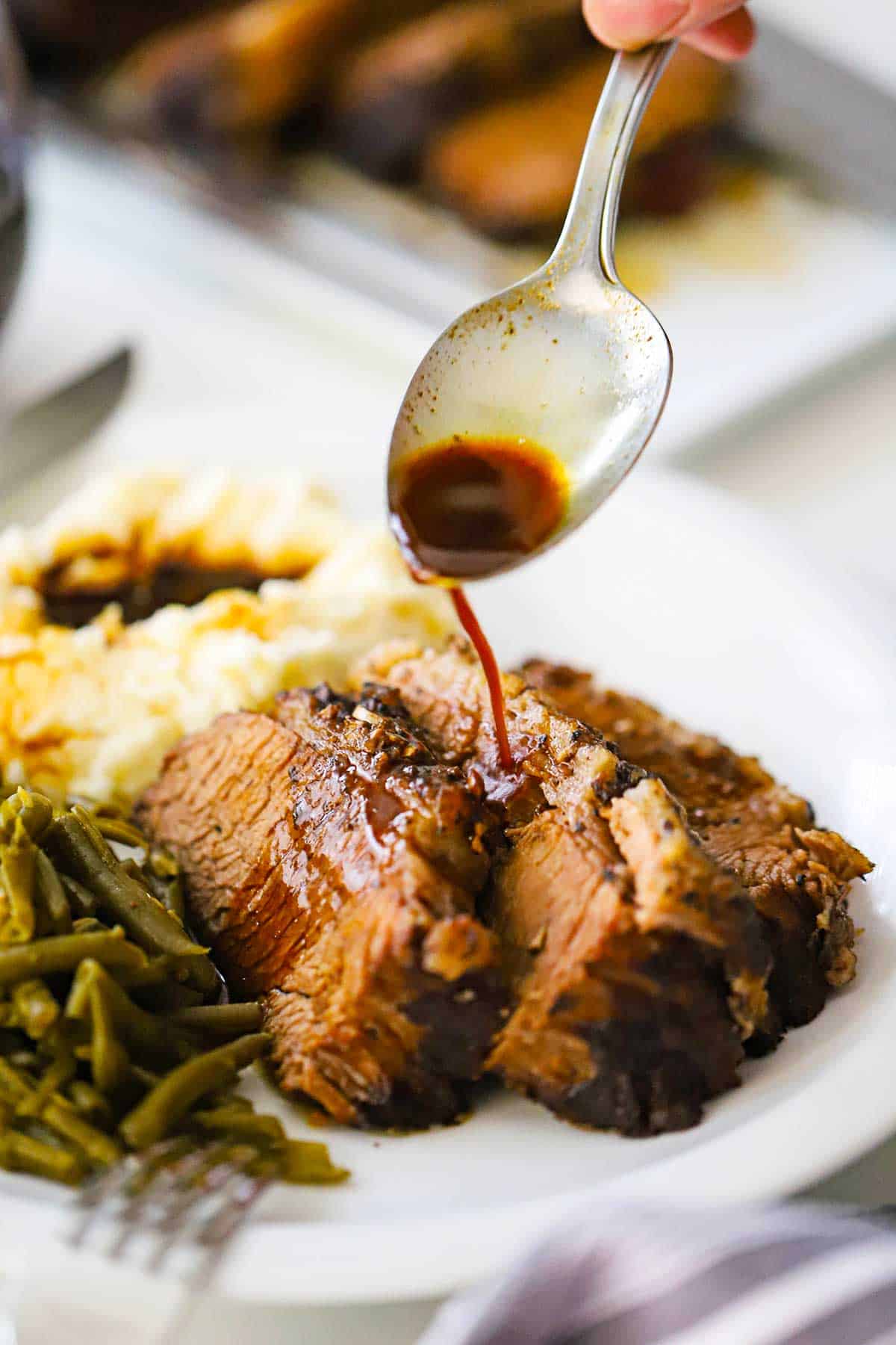 A person pouring an au jus sauce from a spoon onto slices of slow-cooker brisket on a white plate next to a pile of green beans and mashed potatoes.