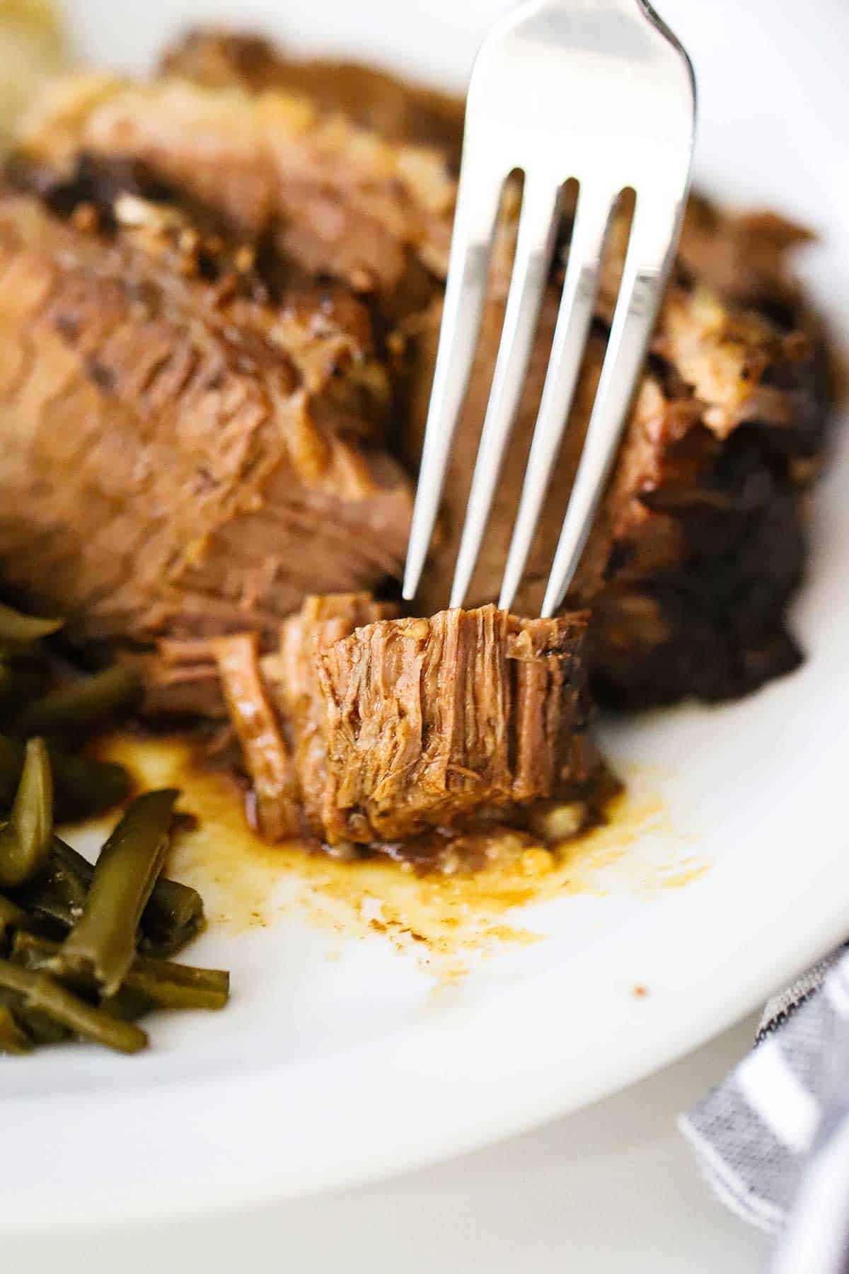 A person using a fork to pierce a piece of slow-cooker brisket that has been pulled away from a large piece of beef on a white dinner plate.