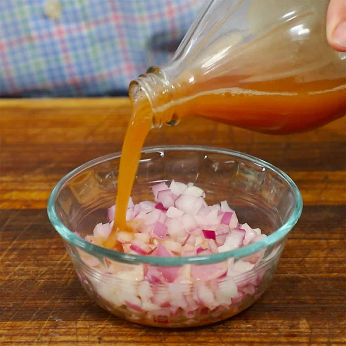 A person pouring apple cider vinegar from a bottle into a small bowl filled with chopped red onion.