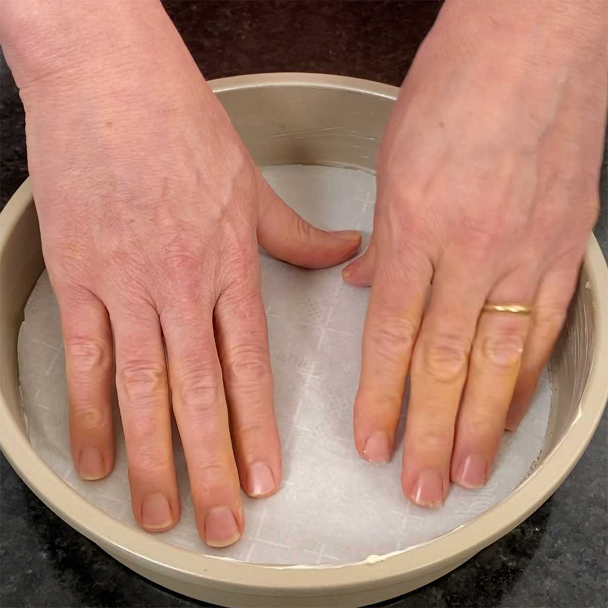 A person using two hands to press a piece of parchment paper into the bottom of a buttered cake pan. 