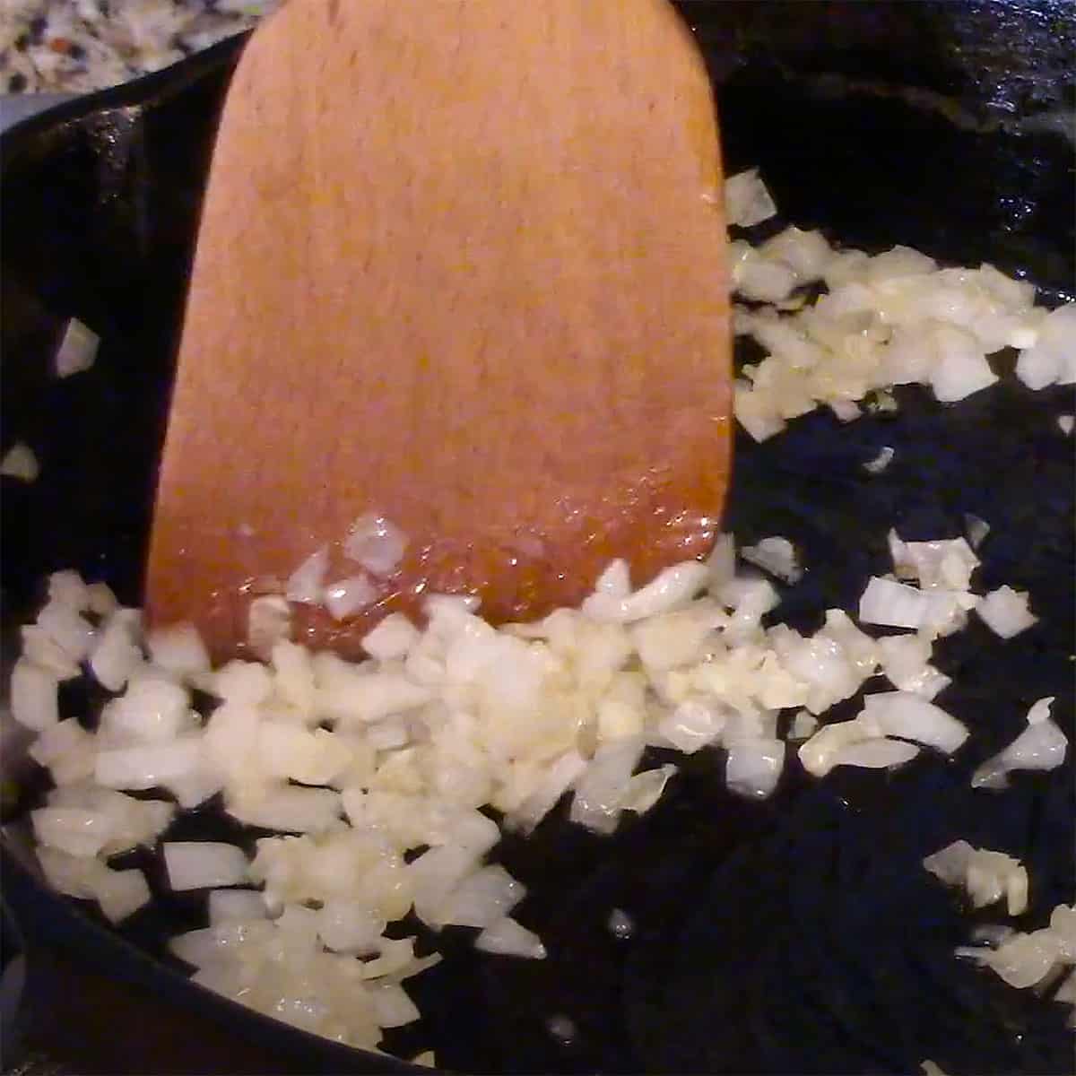 A large wooden spatula stirring chopped onion that is being sautéed in a small cast-iron skillet.
