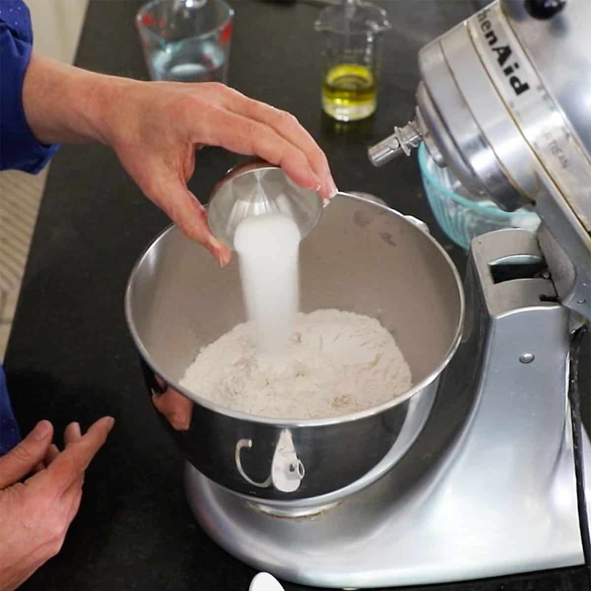 A person transferring sugar into a stand-mixer bowl that is filled with foamy yeast and water.