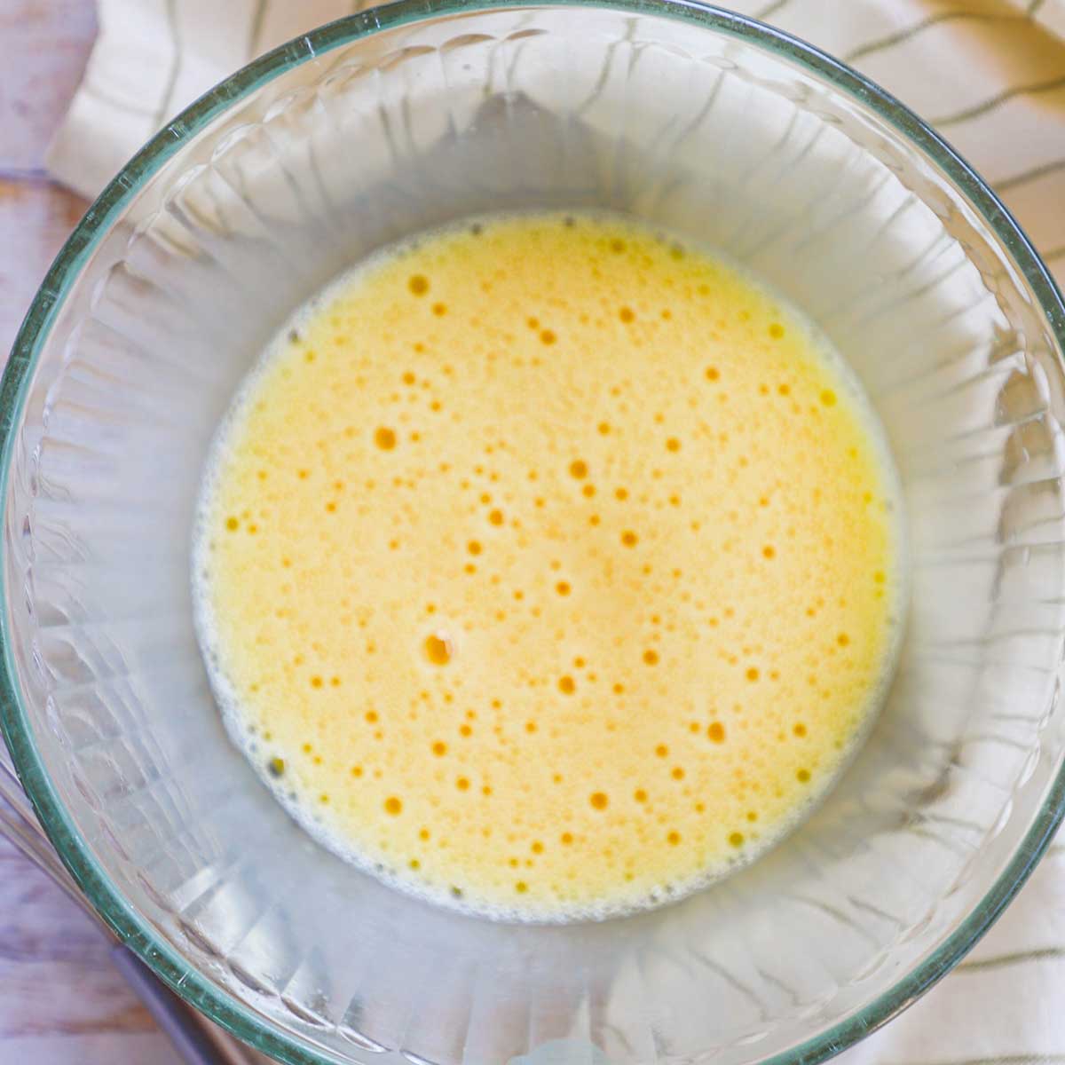 A glass bowl filled with eggs that have been vigorously whisked so that air bubbles are visible on the surface of the eggs. 
