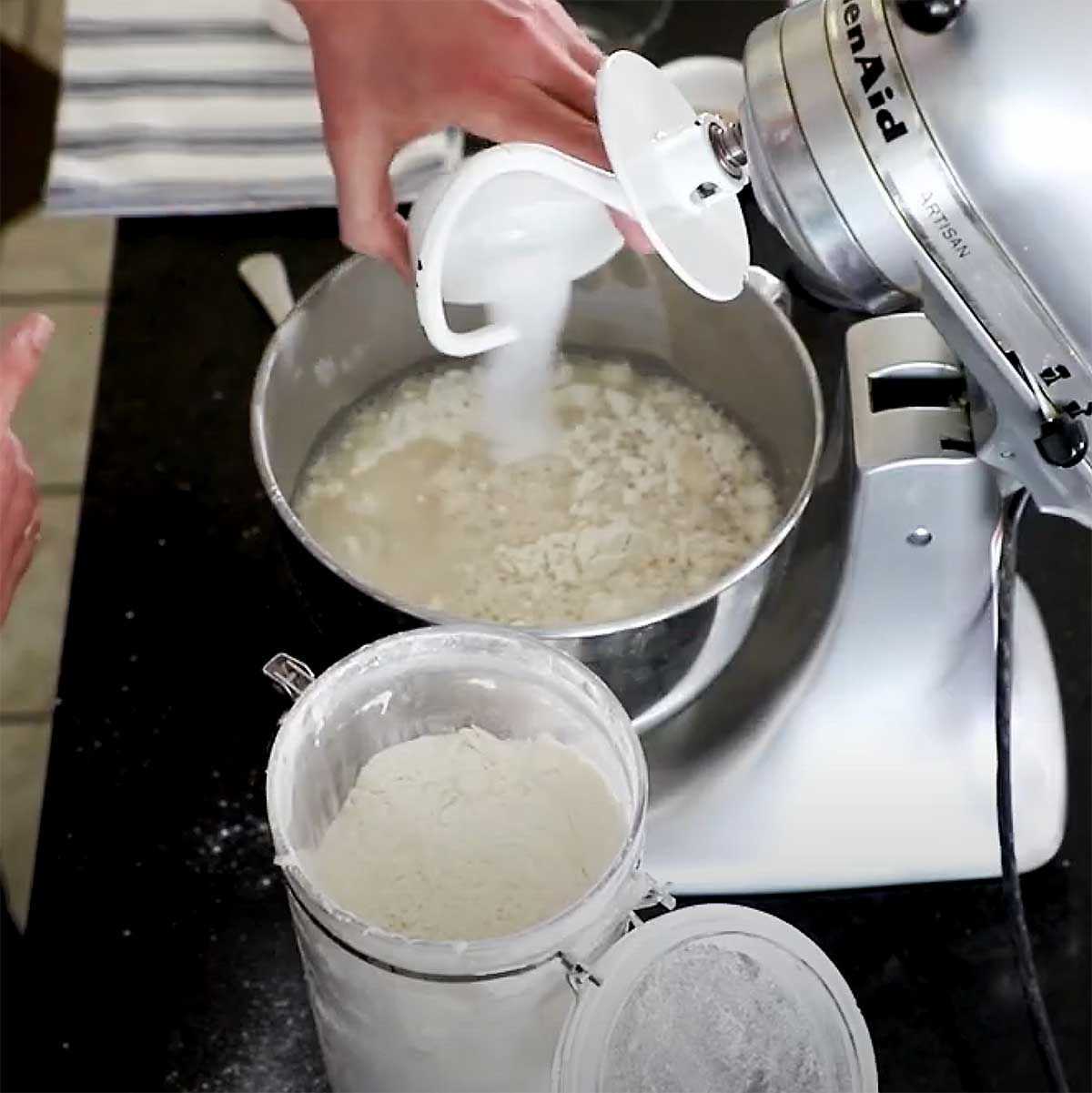 A person adding sugar into the bowl of a mixer that is filled with foamy yeast and water. 