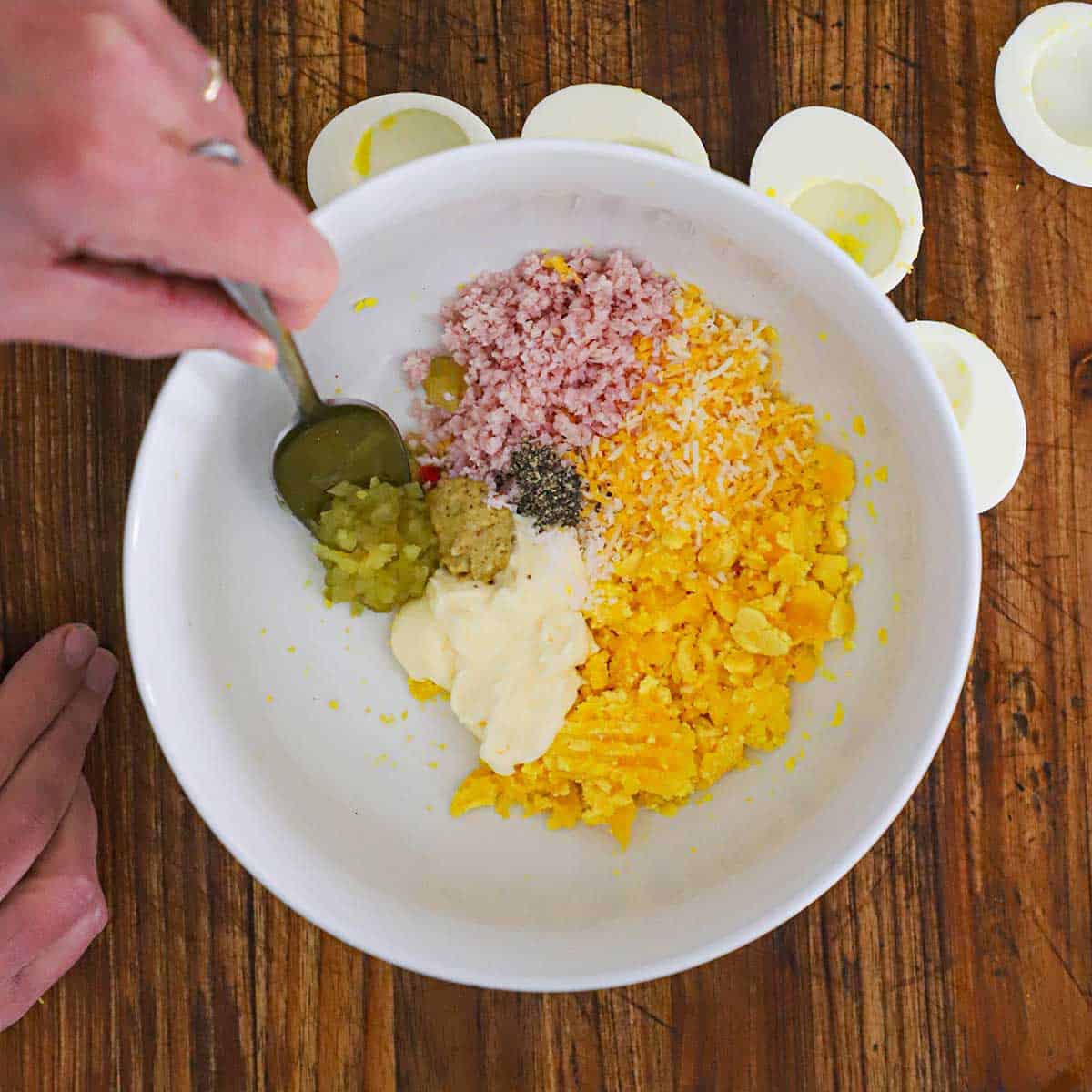A person using a spoon to add pickle relish to a bowl filled with the ingredients for ham and cheese deviled eggs. 