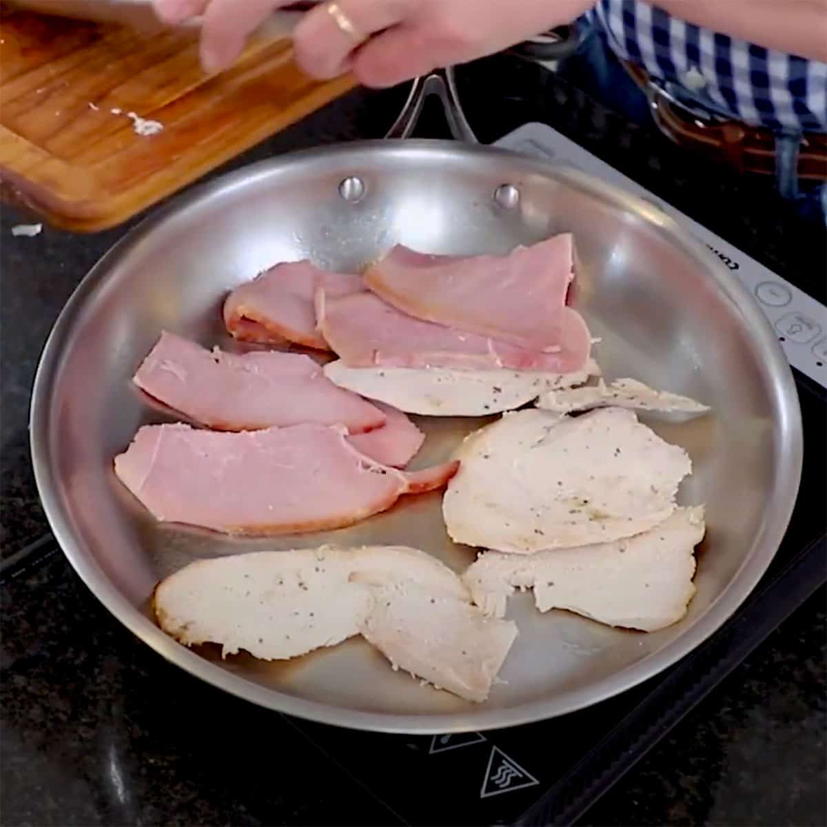Thick slices of ham and turkey being heated in a large silver skillet on a stovetop. 