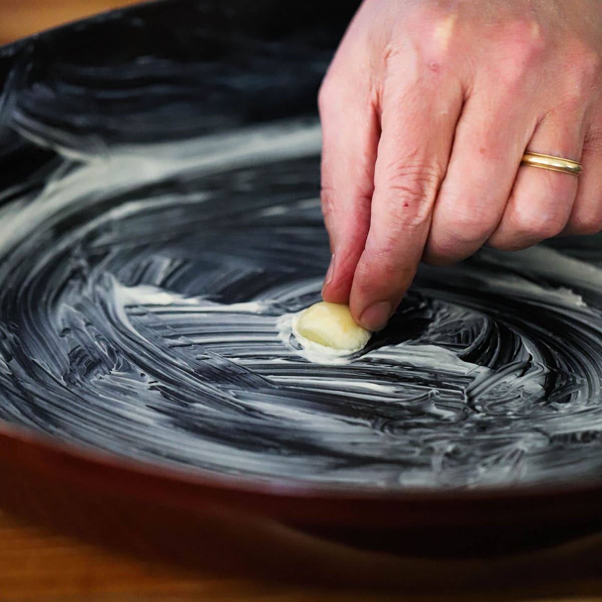 A person rubbing a cut clove of garlic over the base of a buttered oval baking dish. 