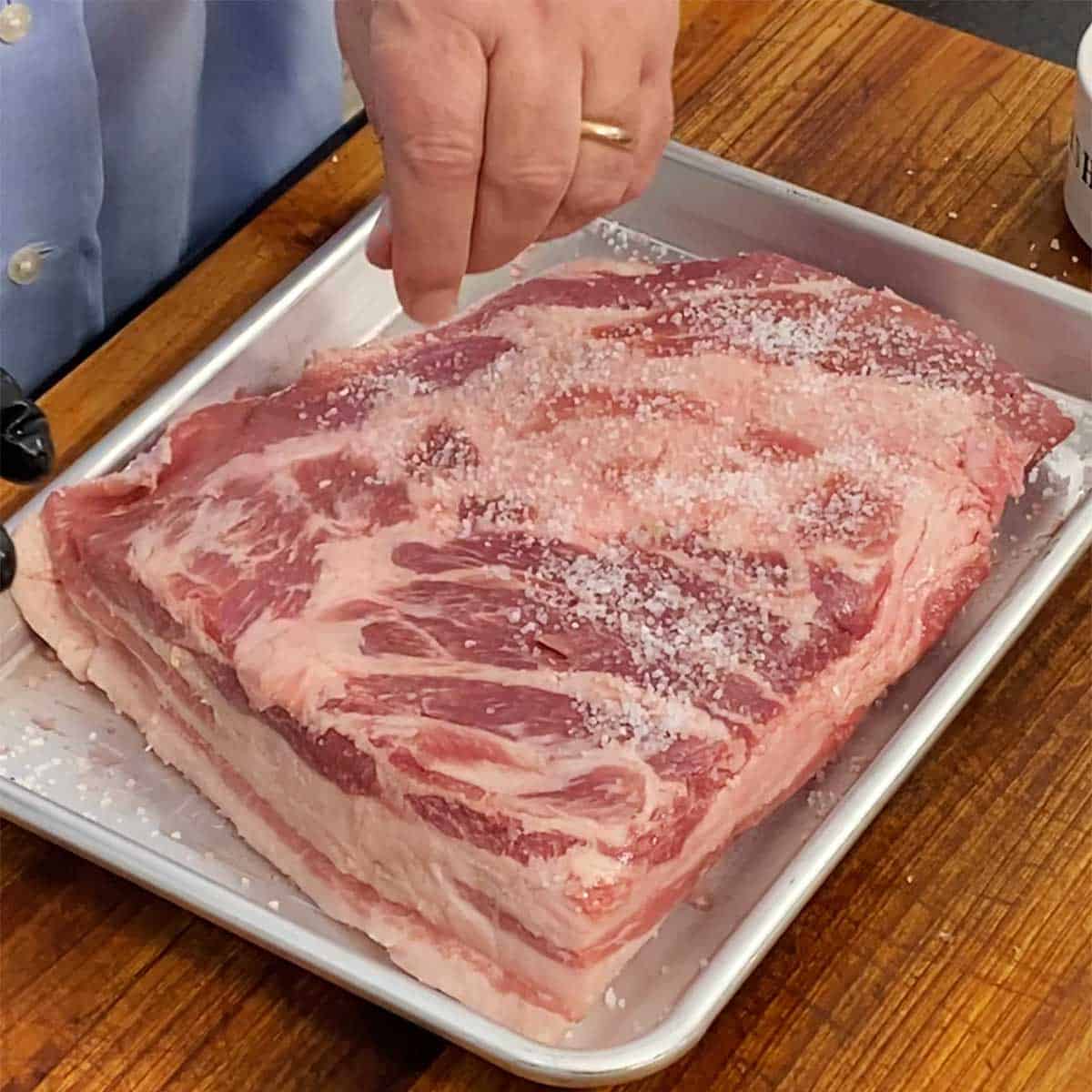 A person sprinkling salt all over an uncooked pork brisket resting in a metal baking pan. 