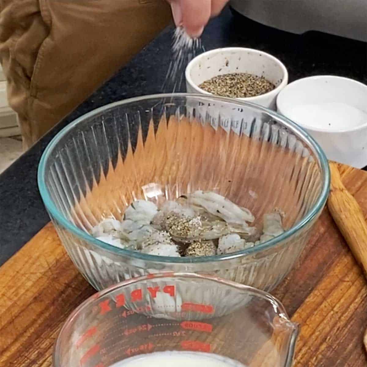 A glass bowl filled with uncooked cleaned shrimp that have been seasoned with salt and pepper. 