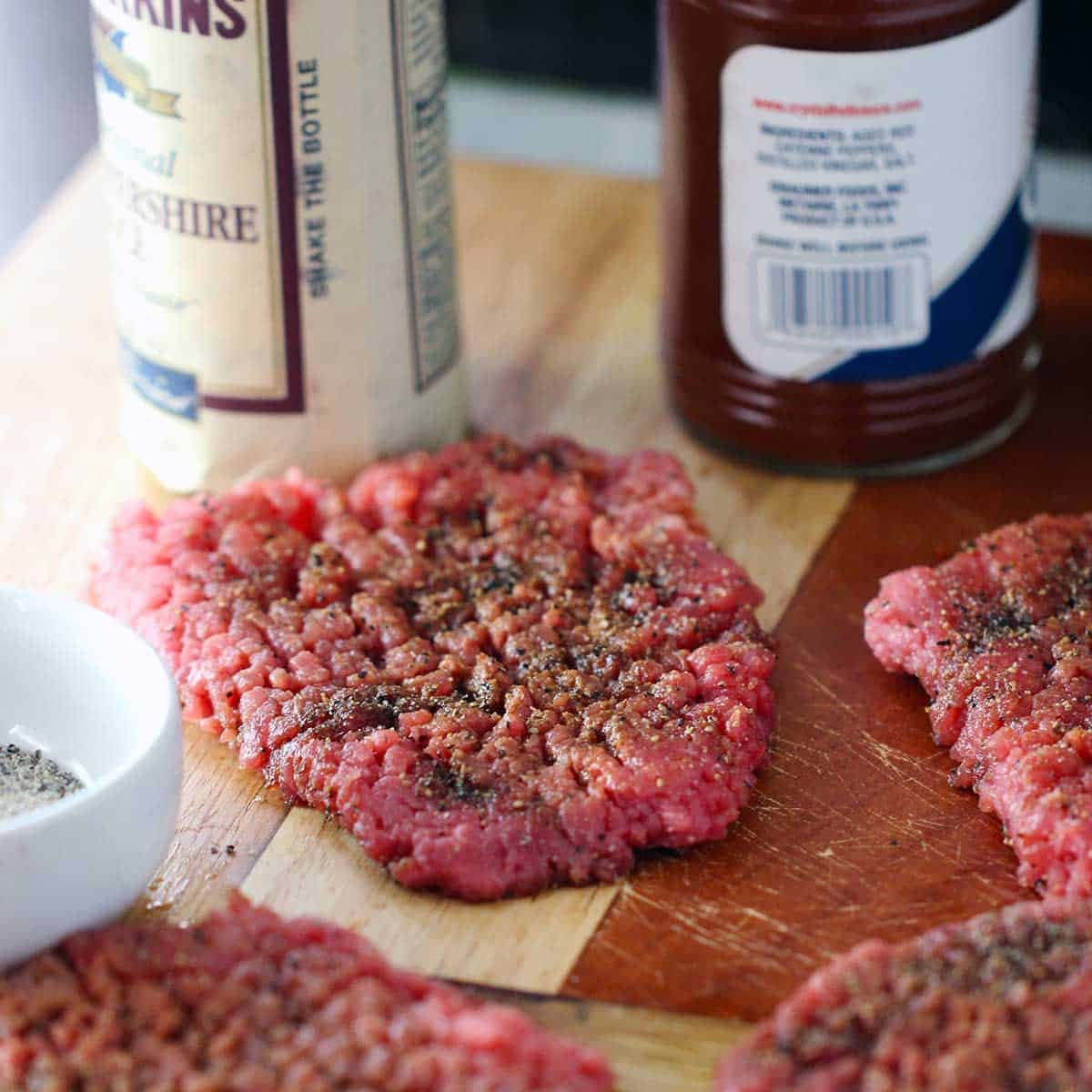 Uncooked tenderized cube steaks that have been seasoned with Worcestershire sauce, hot sauce, and seasoned salt on a cutting board. 