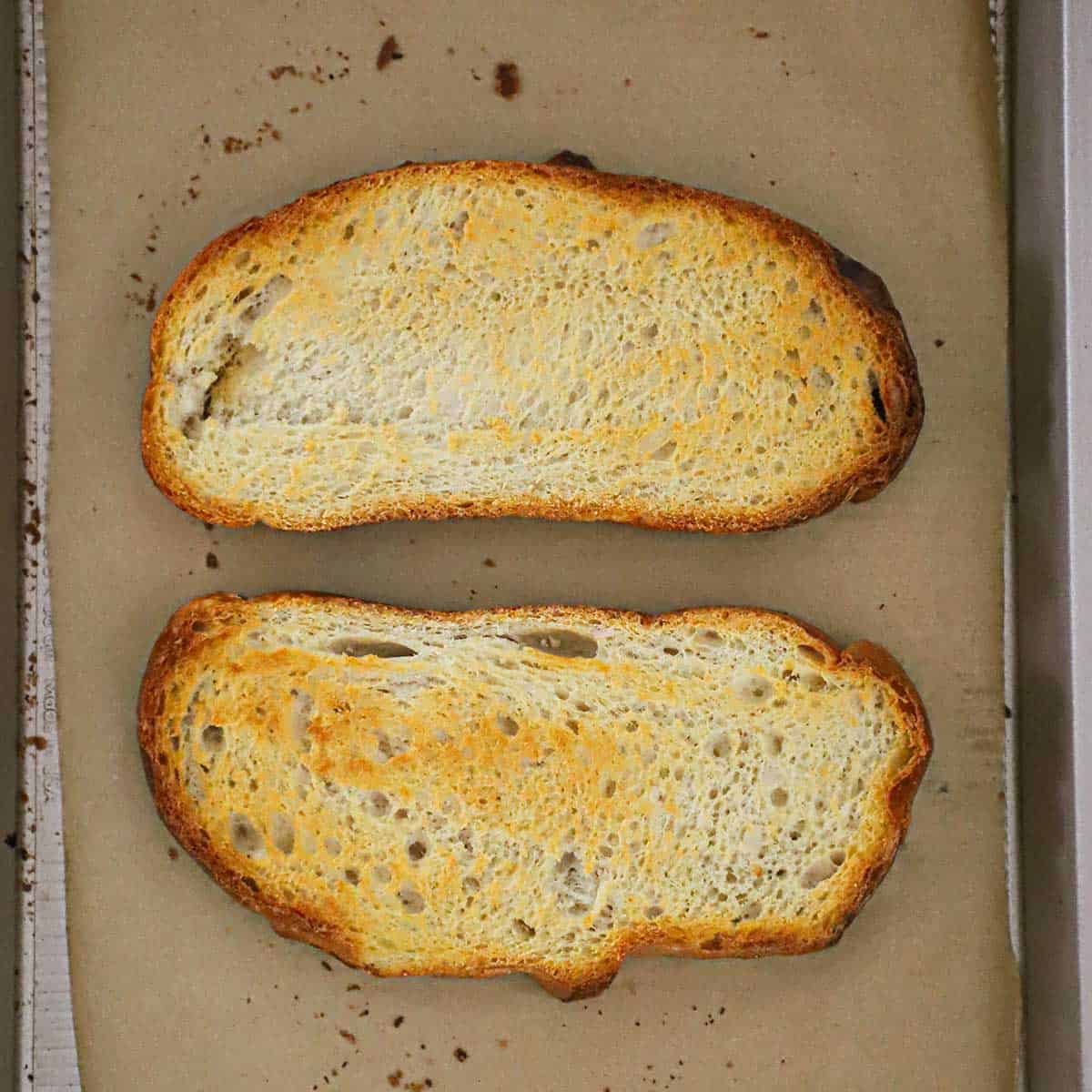 Two toasted slices of sourdough bread on parchment paper in a baking sheet pan. 