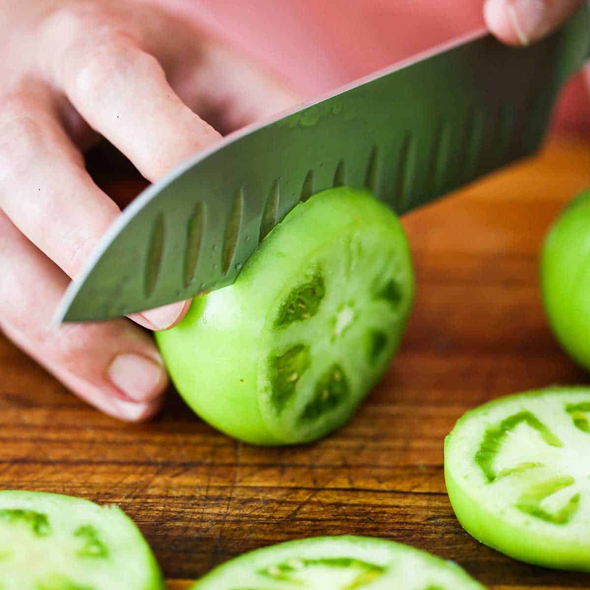 A person using a chef's knife to slice a green tomato on a wooden cutting board. 