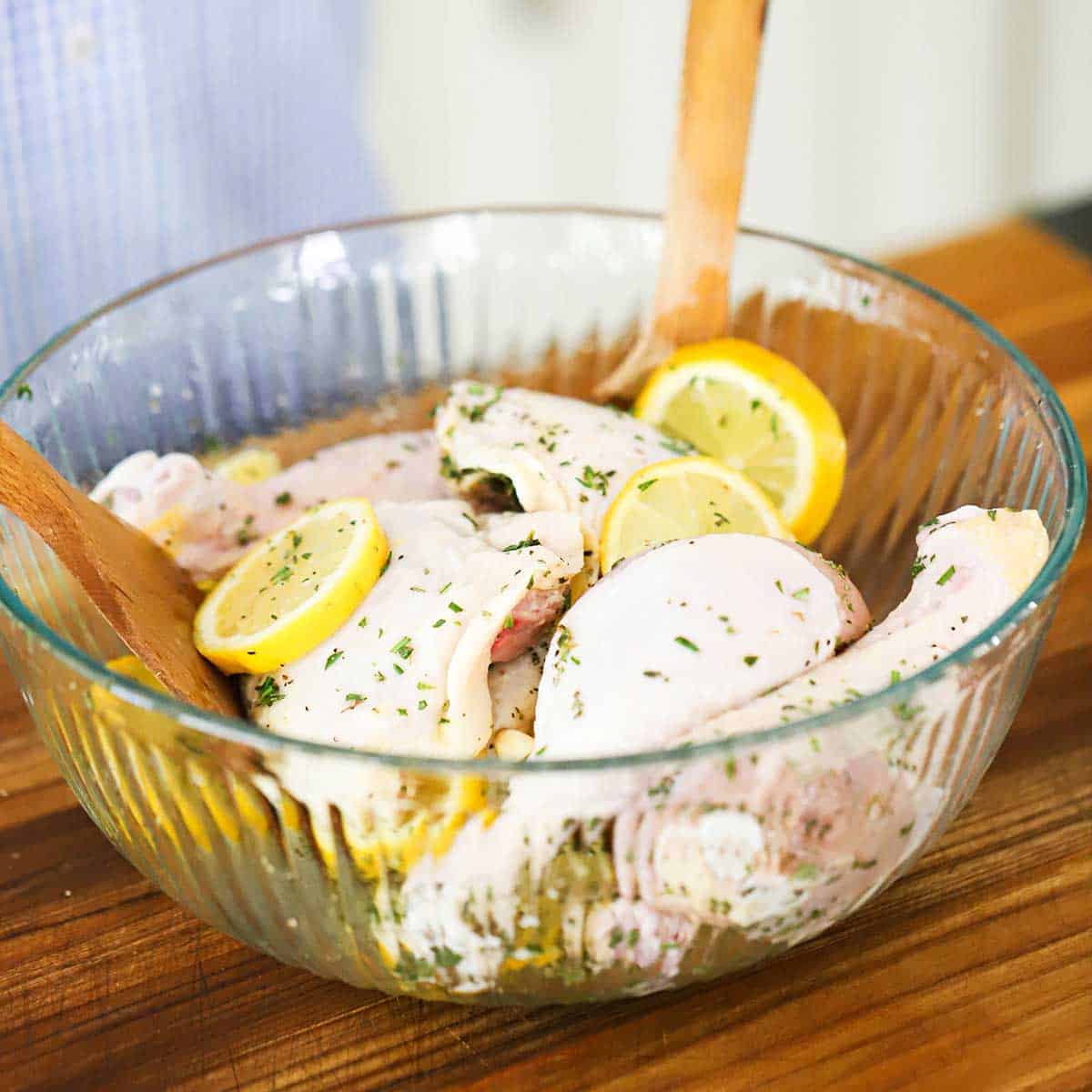 Uncooked chicken pieces in a large glass bowl along with slices of lemon, herbs, and lemon-infused oil.