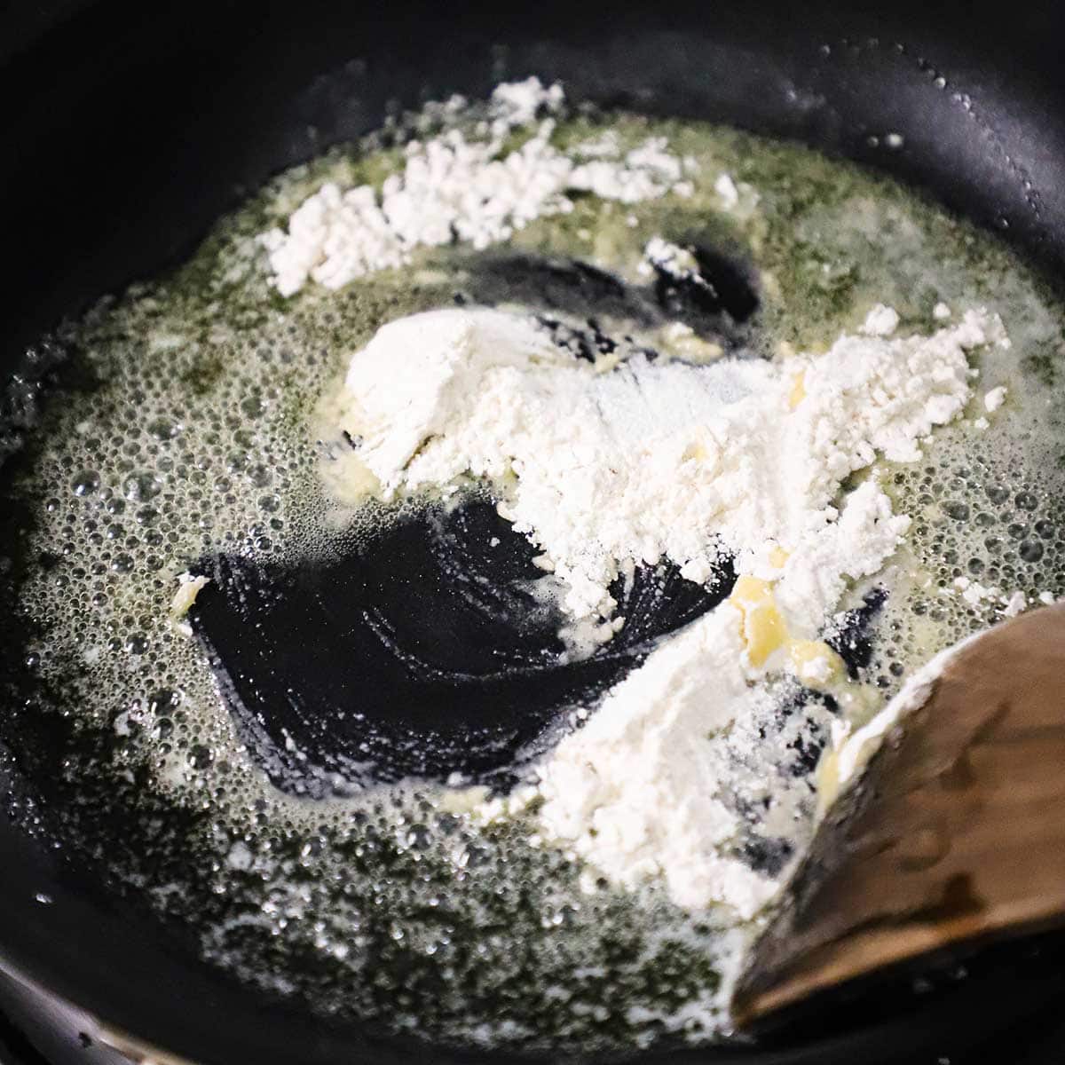 A person using a wooden spatula to stir flour into melted butter in a skillet. 