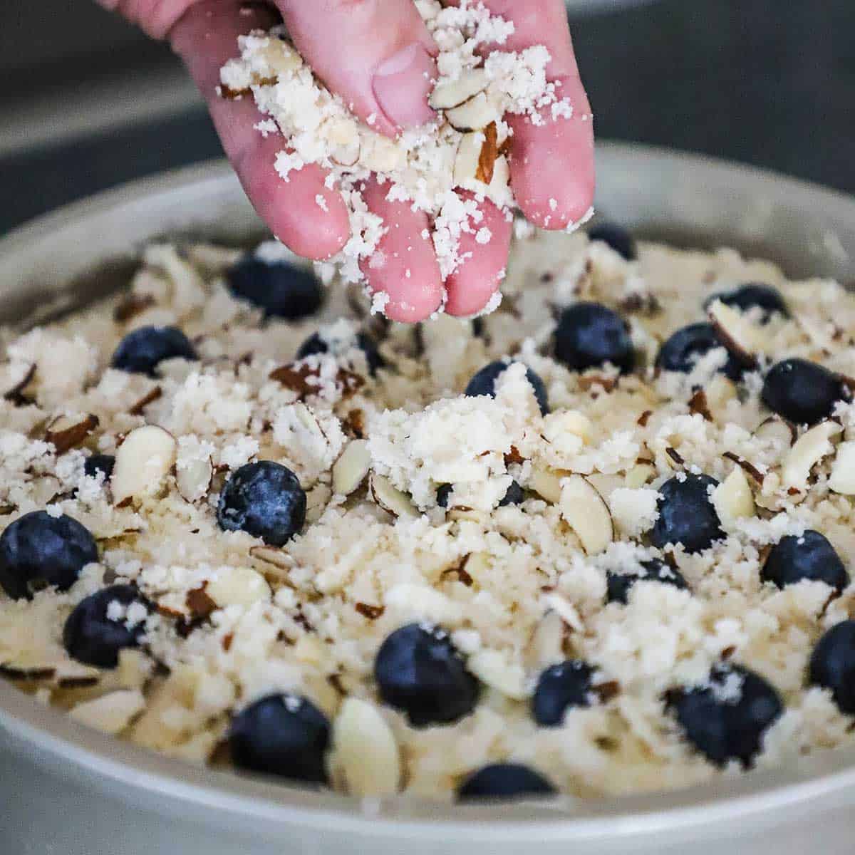 A person sprinkling a streusel topping over blueberries that are resting on the top of an uncooked coffee cake in a metal cake pan.  