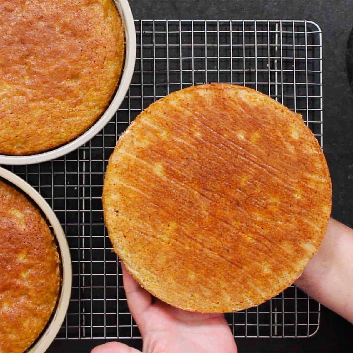 A person placing a carrot cake layer onto a wire baking rack with two pans filled with carrot cake nearby. 