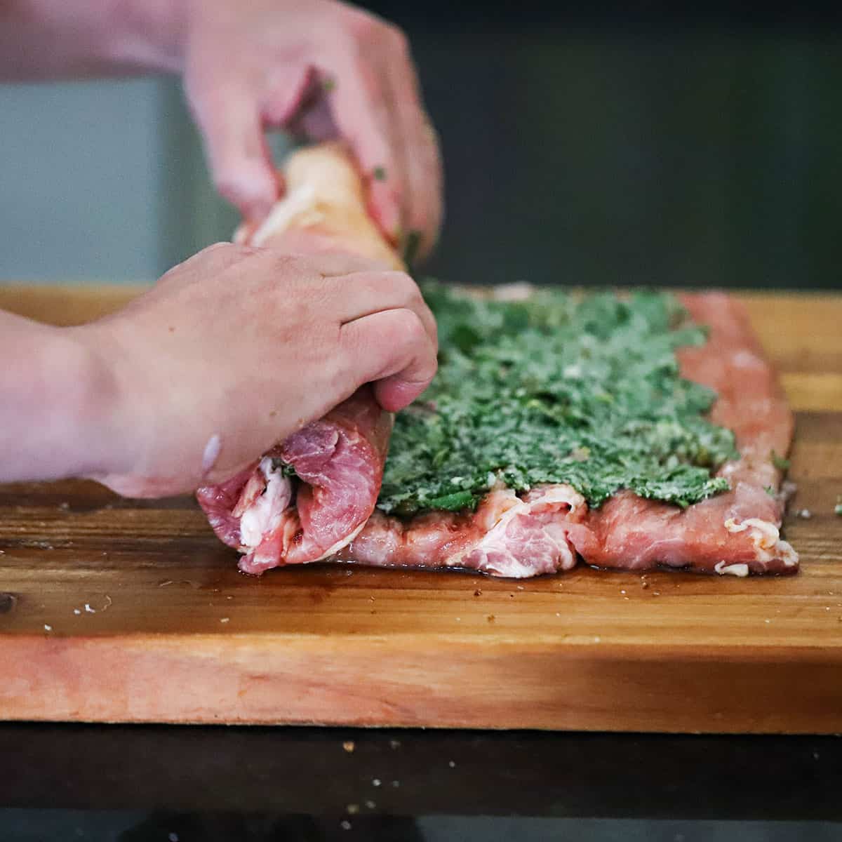 A person rolling up an herb-stuffed pork loin on a cutting board. 