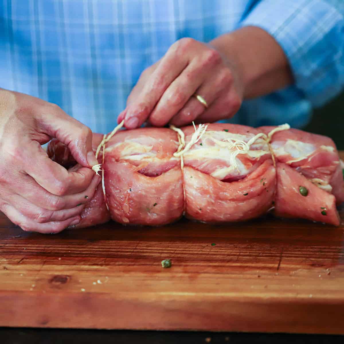 A person using kitchen twine to tie together a rolled up pork loin on a cutting board. 