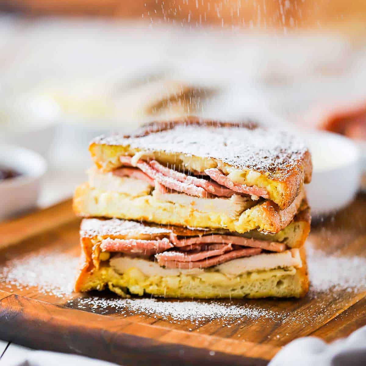Powdered sugar being sprinkled over the tops of a Monte Cristo sandwich that has been cut in half and placed on top of each other. 