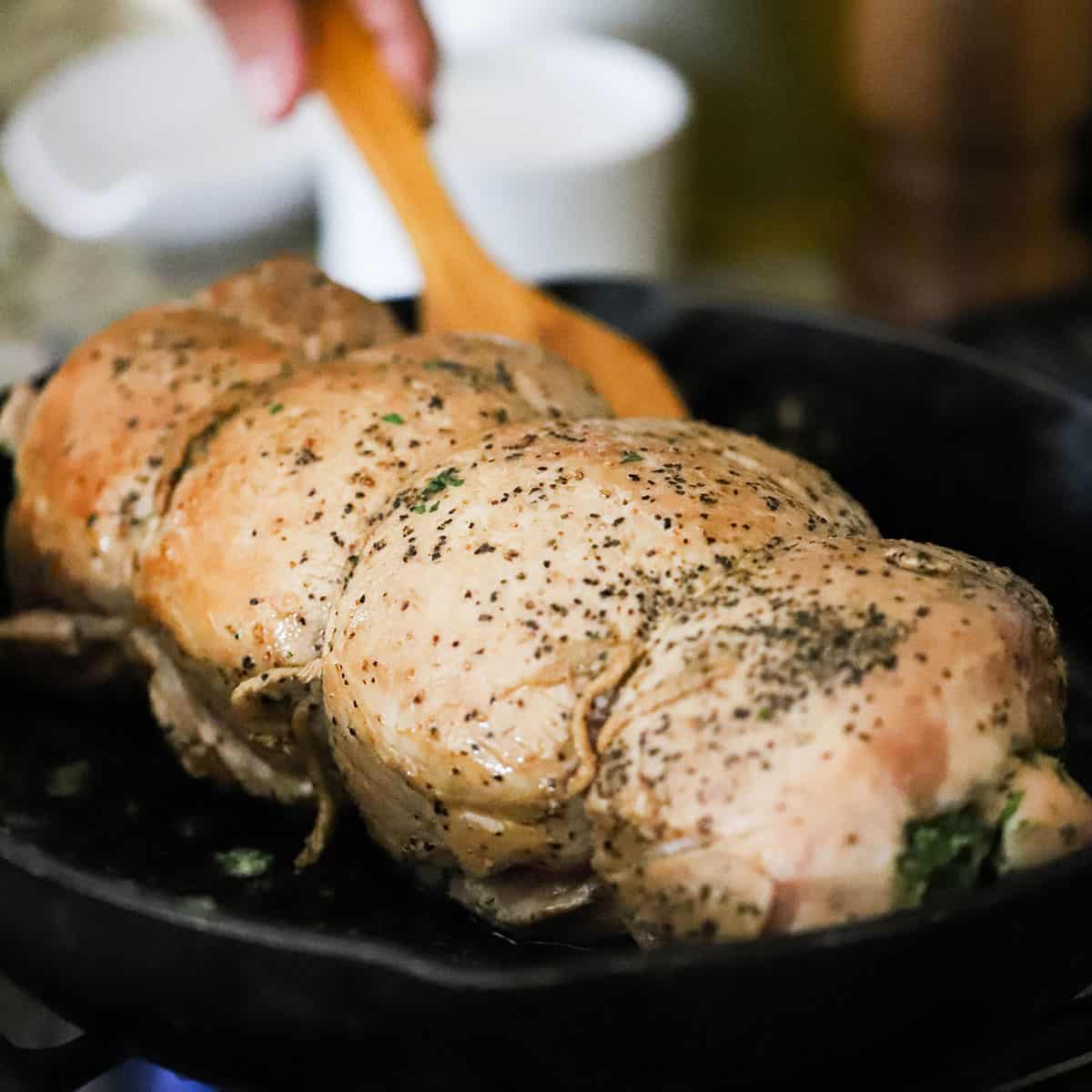 An herb-stuffed pork loin that is tied together with string being seared in a large cast-iron skillet. 