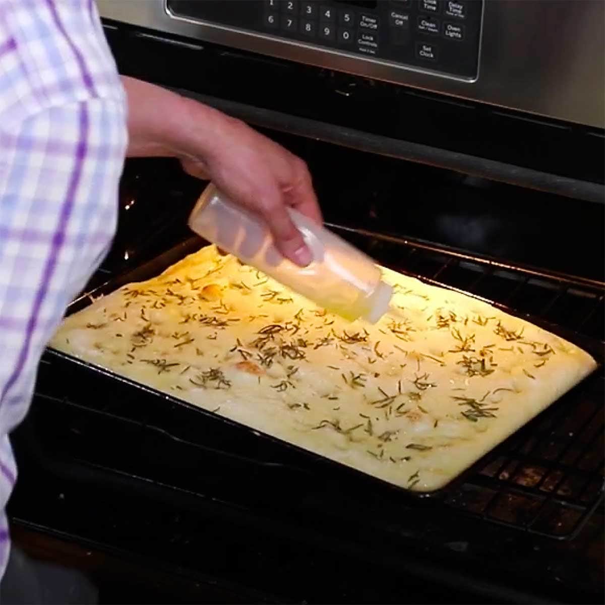 A person squirting olive oil from a plastic squirt bottle onto focaccia dough that is baking in an oven. 