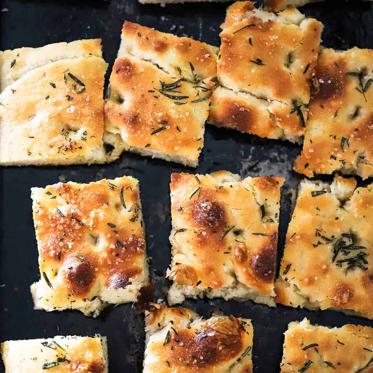 Focaccia bread with rosemary and olive oil cut into squares on a black baking sheet pan. 