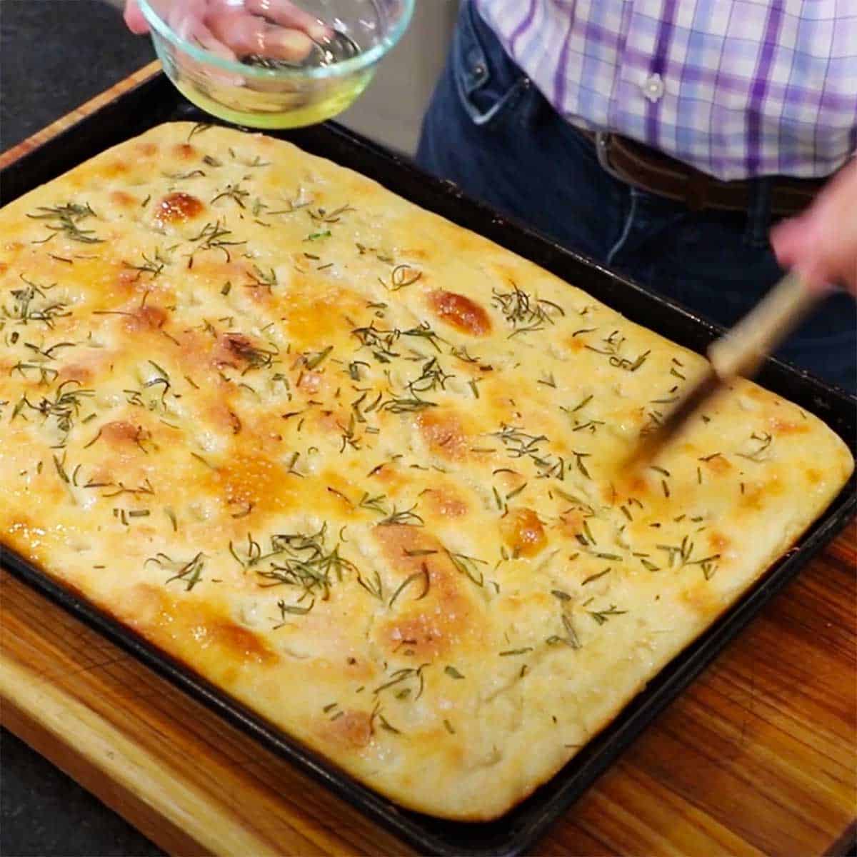 A person brushing olive oil over the top of freshly baked focaccia bread with rosemary in a baking sheet on a cutting board. 
