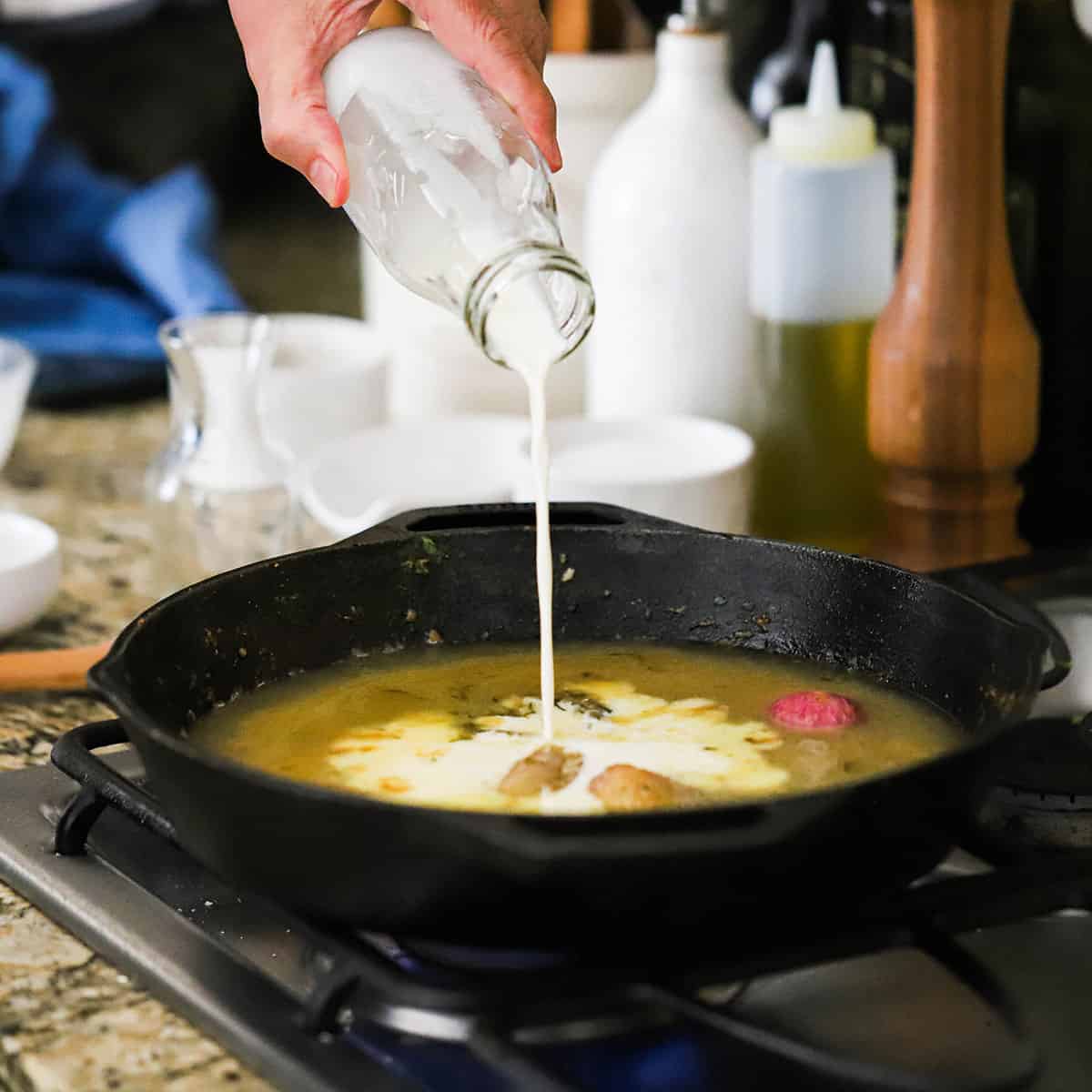 A person pouring cream into a skillet of simmering sauce of white wine, shallots, and garlic. 