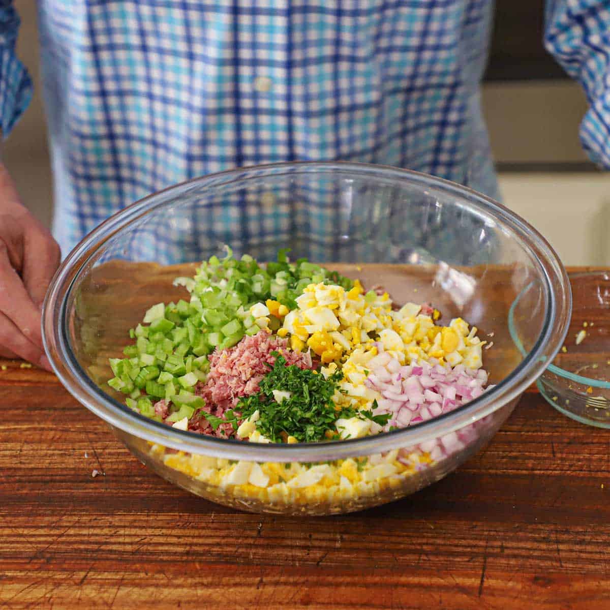 A person standing behind a glass bowl filled with the un-mixed ingredients of ham salad on a wooden cutting board. 