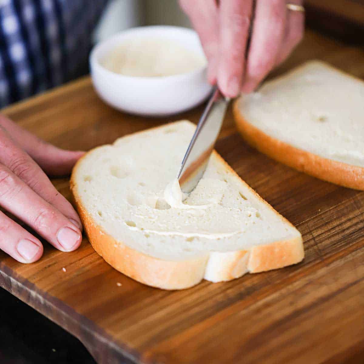 A person using a butter knife to spread a mayonnaise and Dijon mustard mixture onto a slice of sourdough bread. 