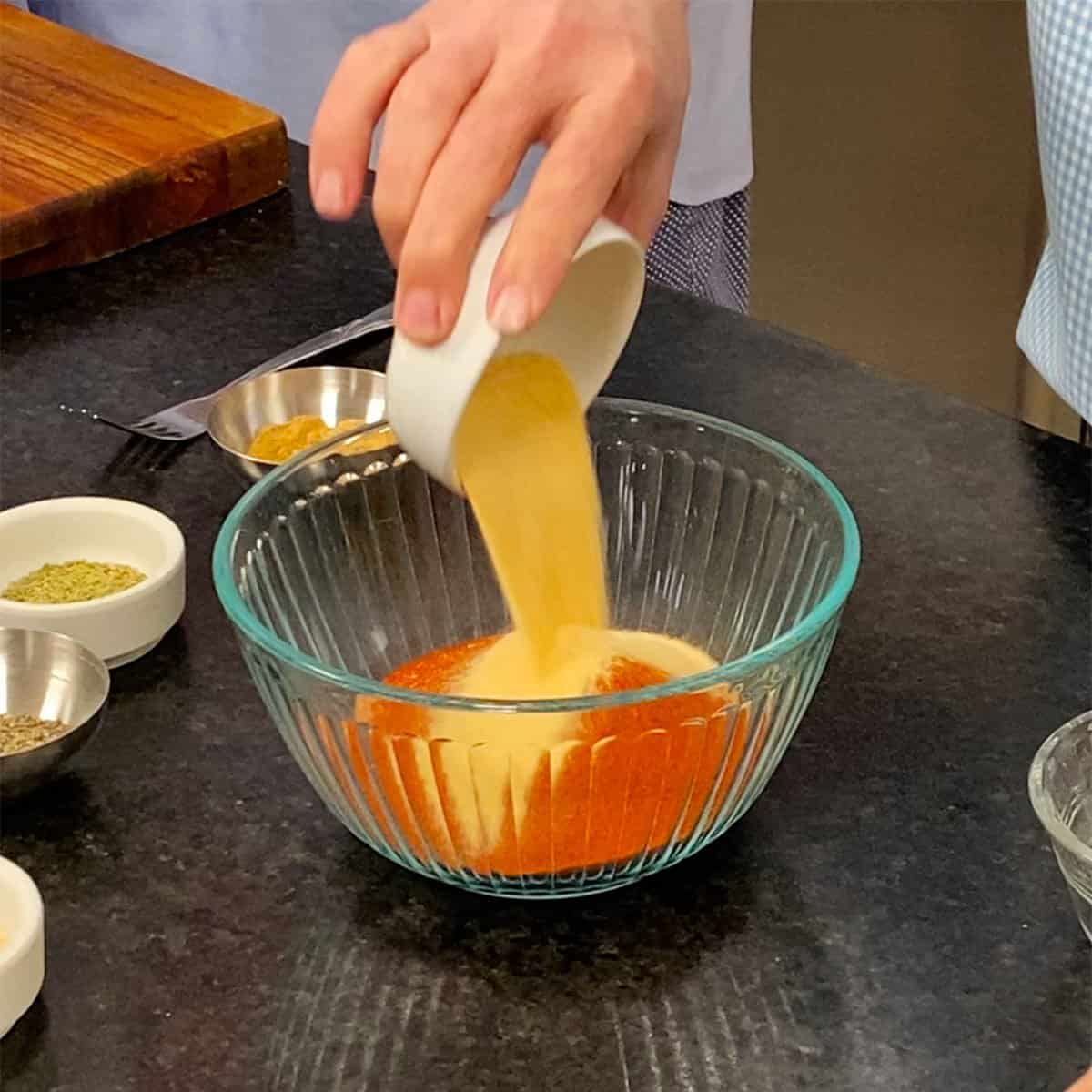 A person dumping garlic powder into a glass bowl that contains onion powder, paprika, brown sugar, and granulated sugar.