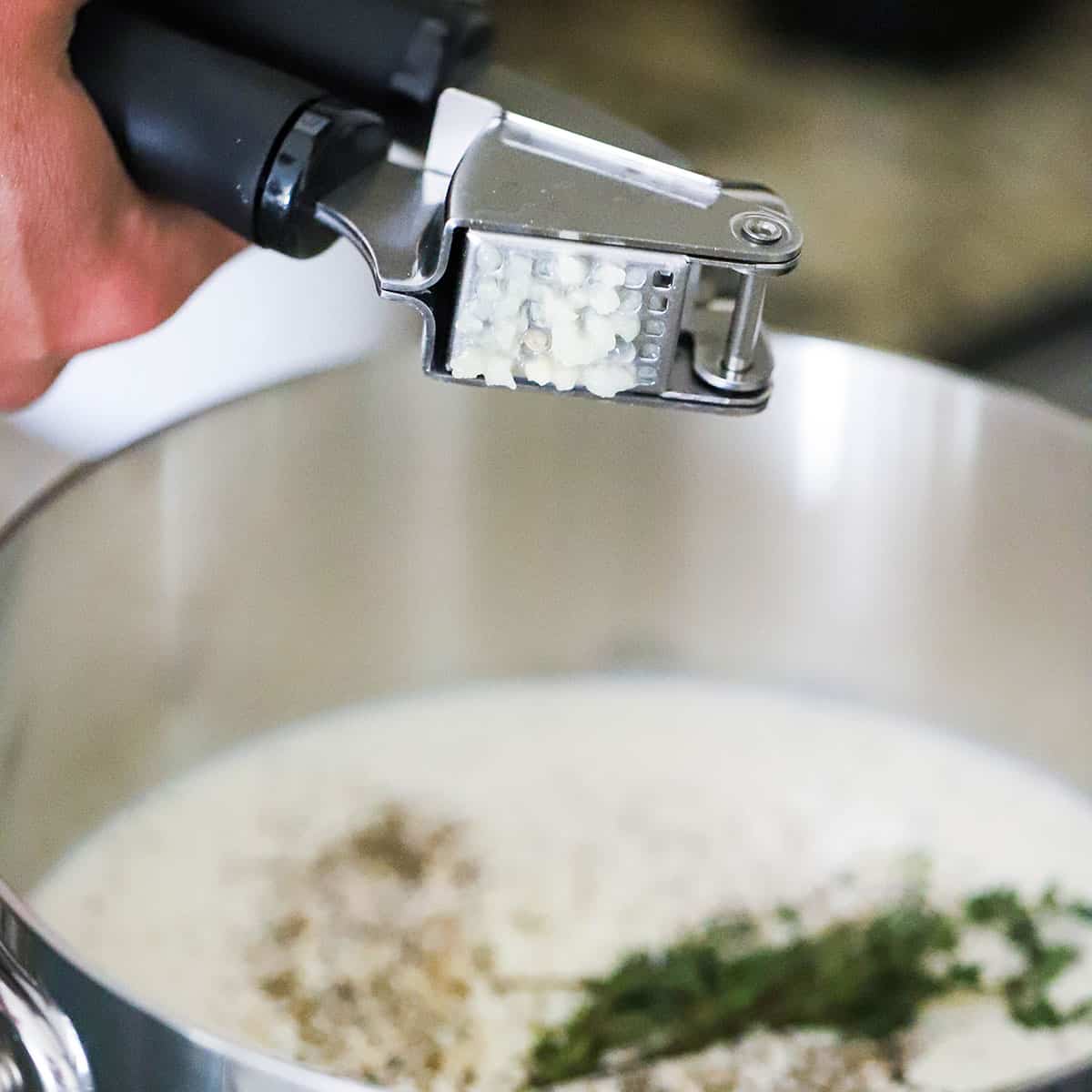 A person using a garlic press to add crush garlic to a saucepan filled with cream, pepper, and herbs. 