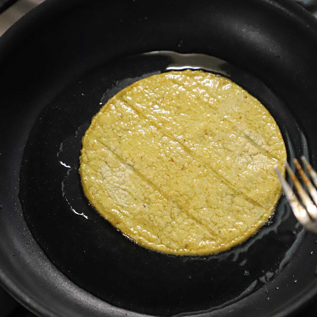 A corn tortilla being lightly fried in vegetable oil in a small black skillet. 
