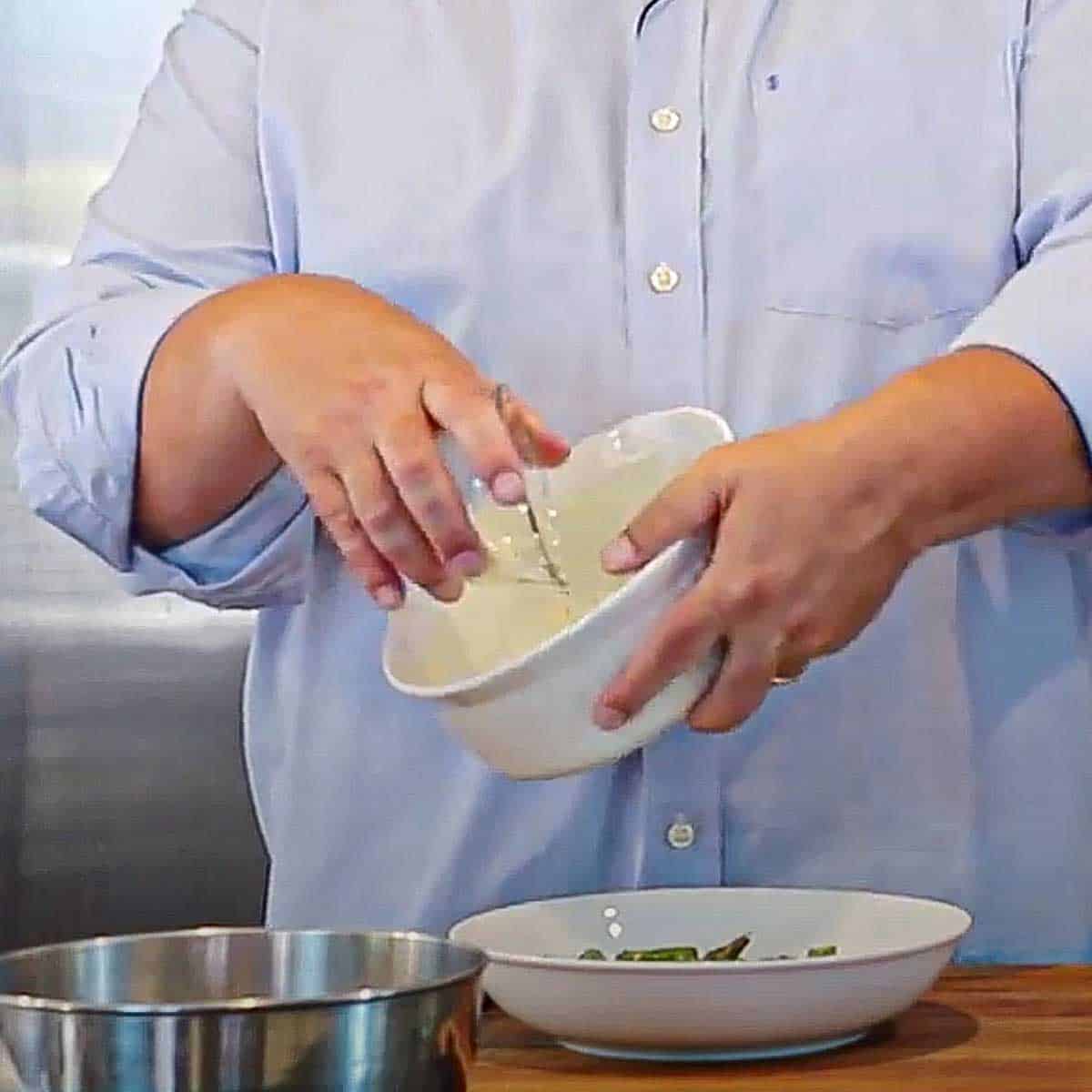 A person dumping Parmesan cheese from a small glass bowl into a large white bowl that has been greased with softened butter.