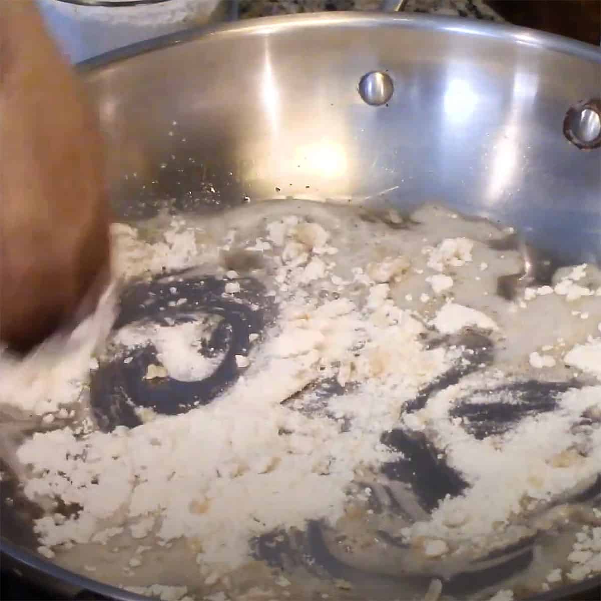 A person using a wooden spoon the incorporate flour into melted bacon grease in a large skillet. 