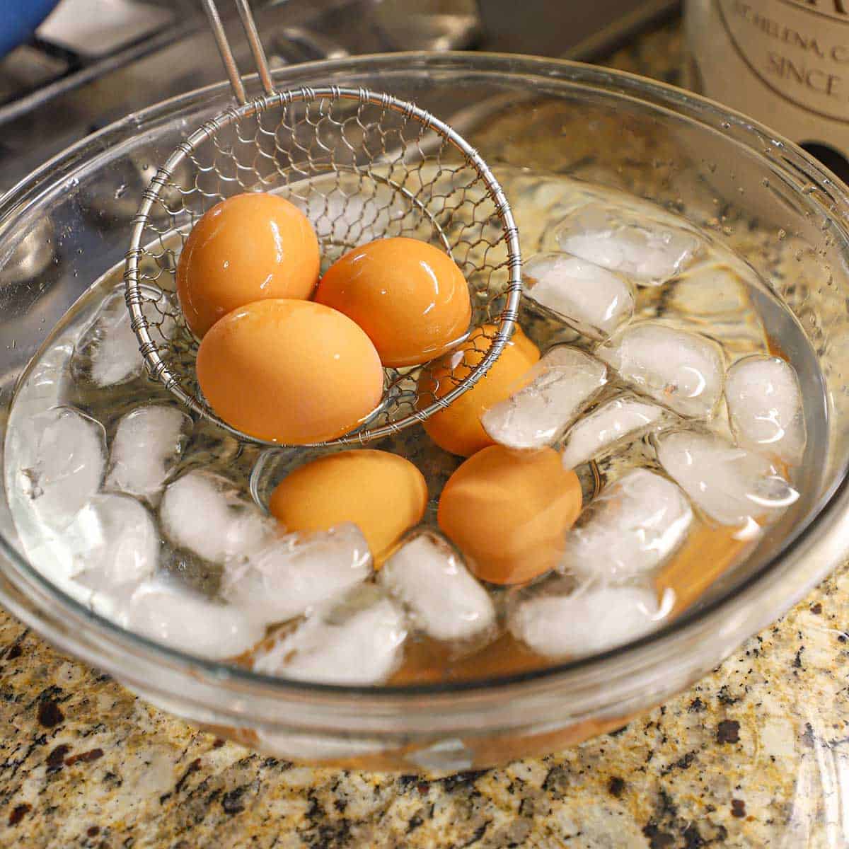 A person using a metal spider to place hard boiled eggs into an ice bath in a glass bowl. 