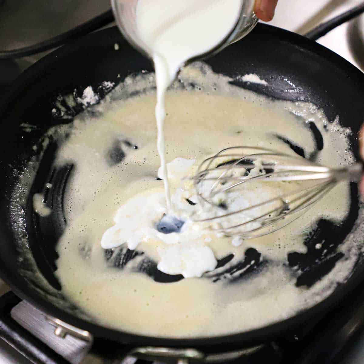 A person whisking milk into a skillet that has a flour and butter roux in it. 