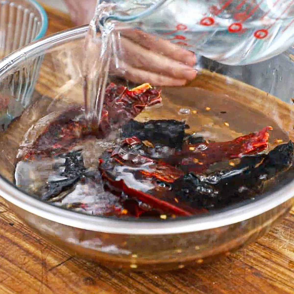 A person pouring hot tap water from a large glass measuring cup into a large glass bowl filled with dried chiles.
