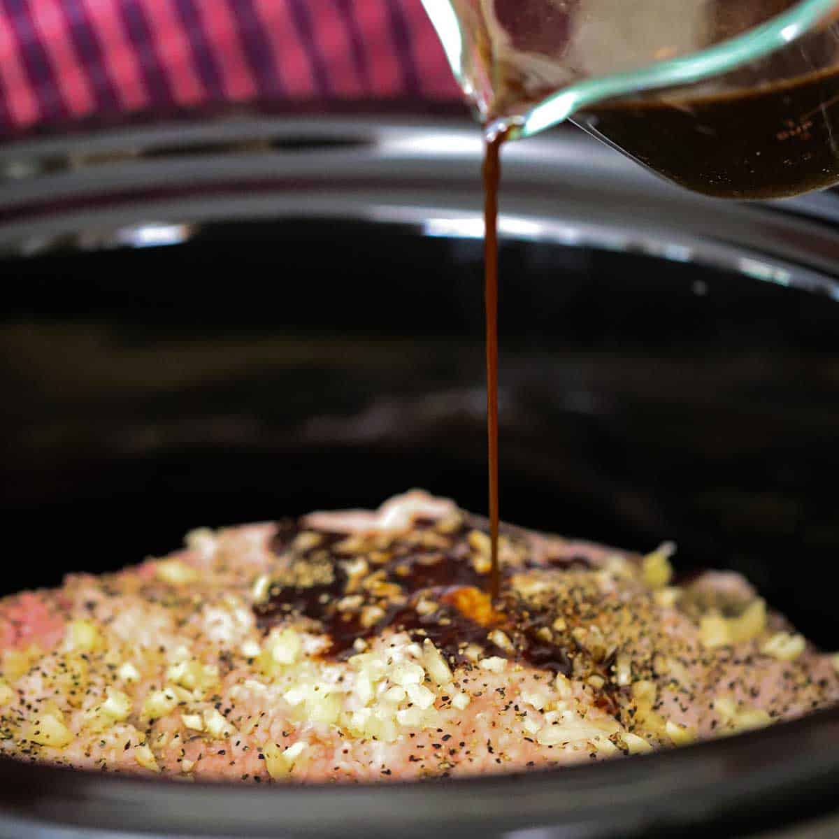 A person pouring a soy sauce and Worcestershire sauce mixture from a glass measuring cup onto an uncooked brisket topped with minced garlic and is resting the a slow cooker.