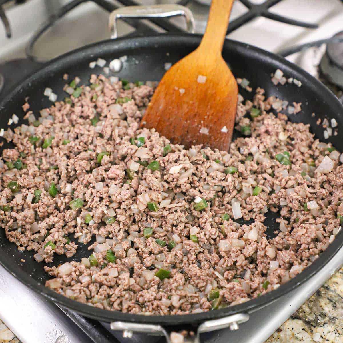 Ground beef being cooked with chopped onions and jalapeños in a large skillet. 