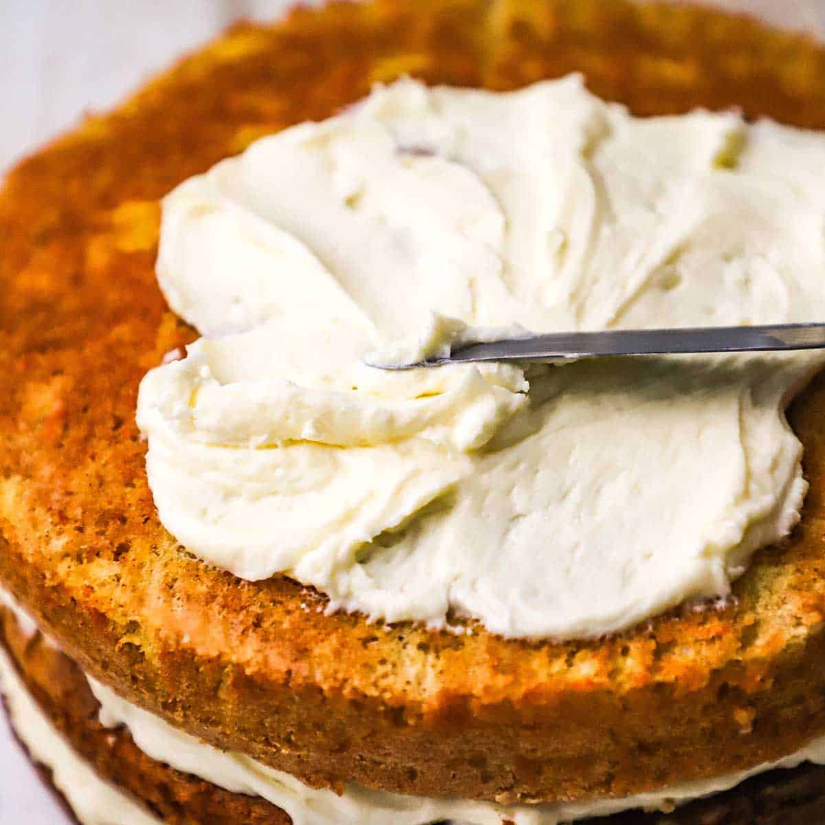 A person using an offset spatula to spread cream cheese icing over the top of a three-layer carrot cake. 