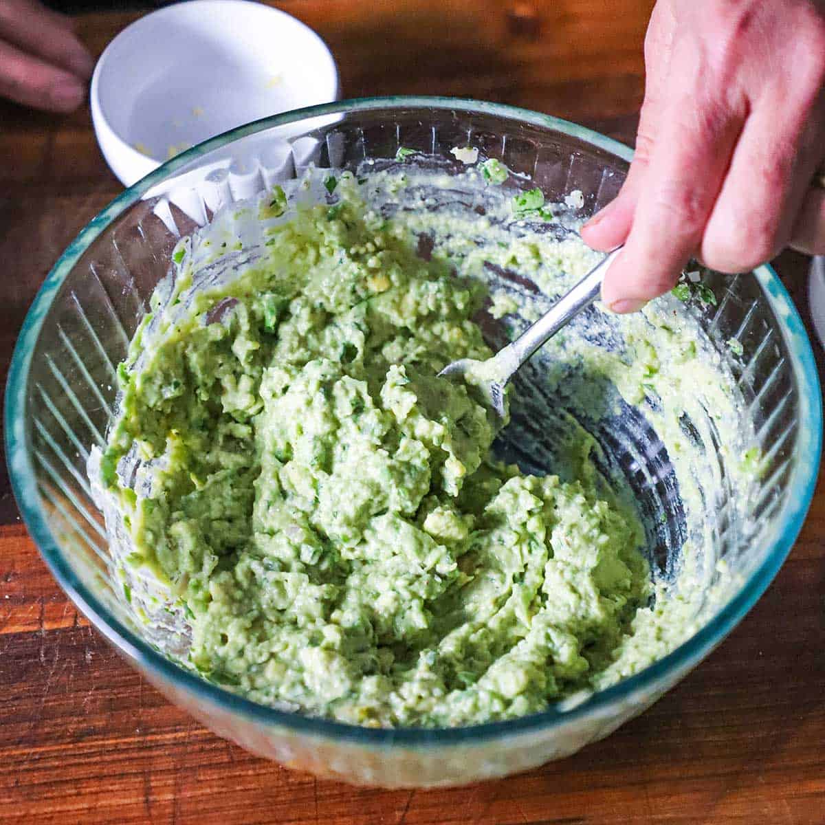 A person using the back of a fork to mash egg yolks, chunks of avocado, onion, and herbs in glass bowl. 