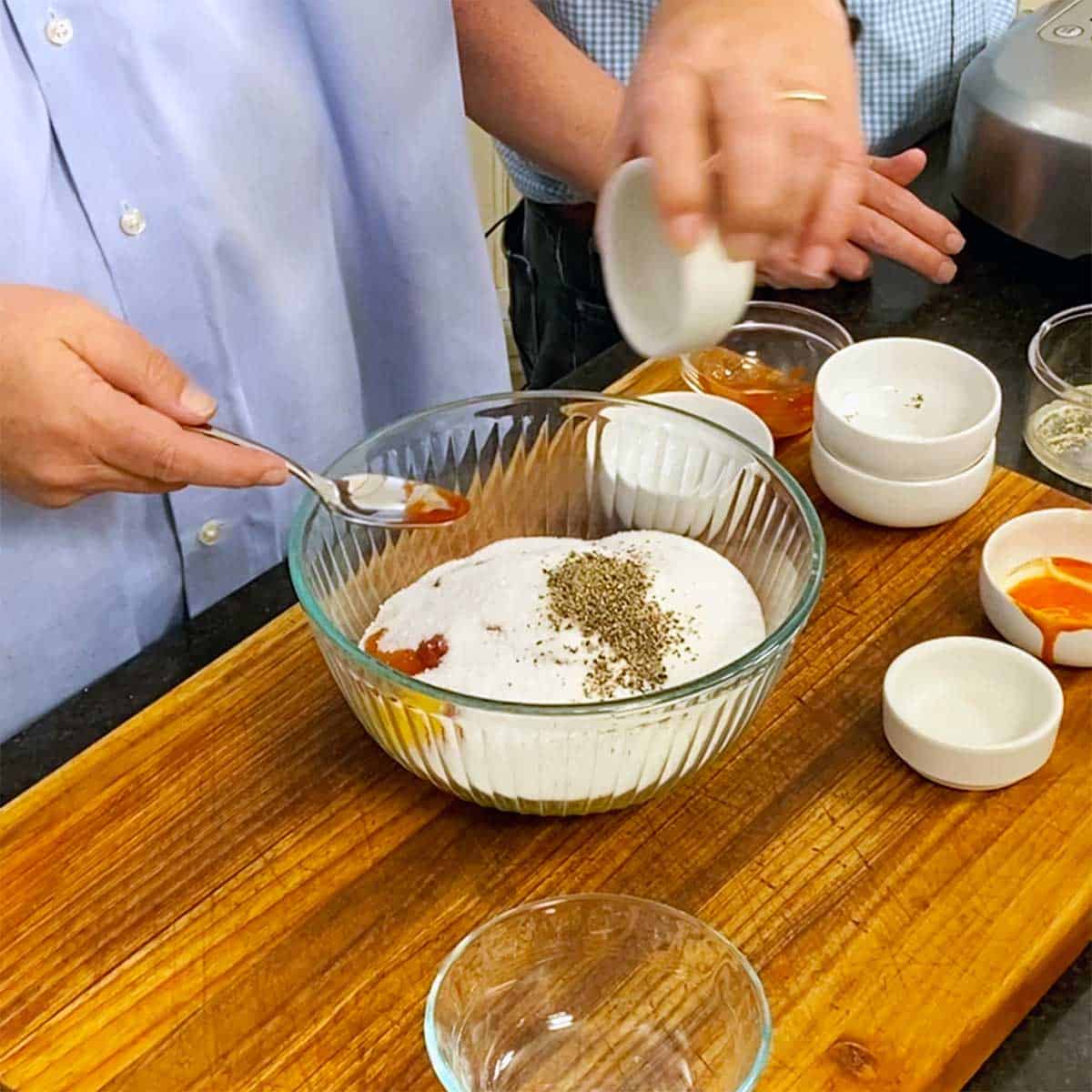 A person transferring salt from a small bowl into a larger bowl filled with spices, ketchup, and hot sauce.