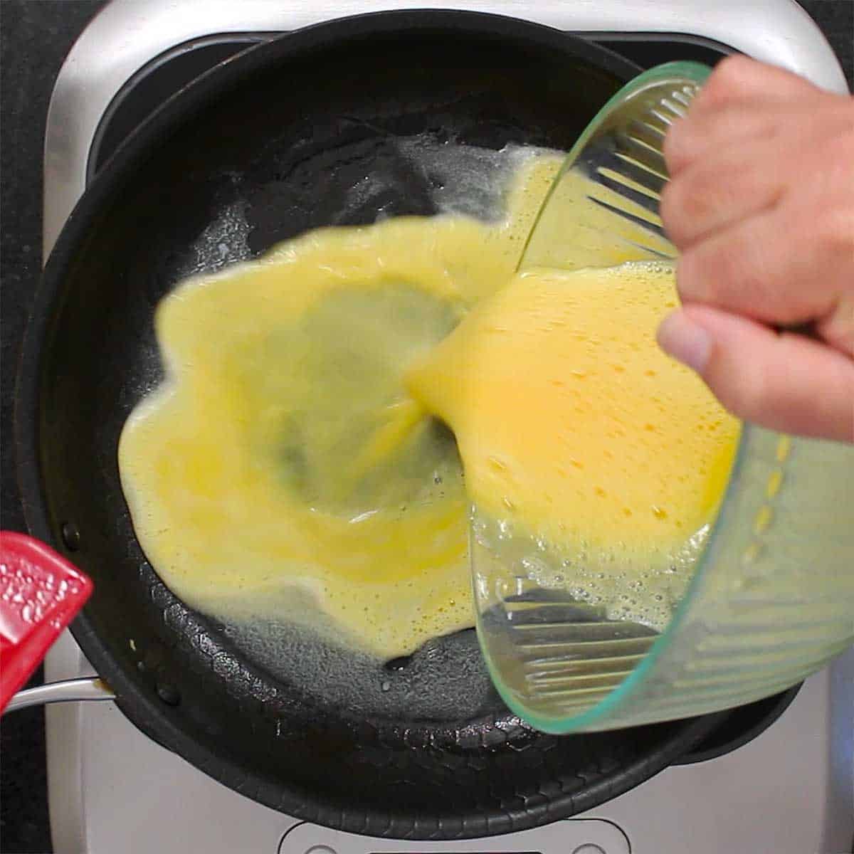 A person pouring whisked whole eggs into a skillet with a thin layer of melted butter in the bottom of it. 
