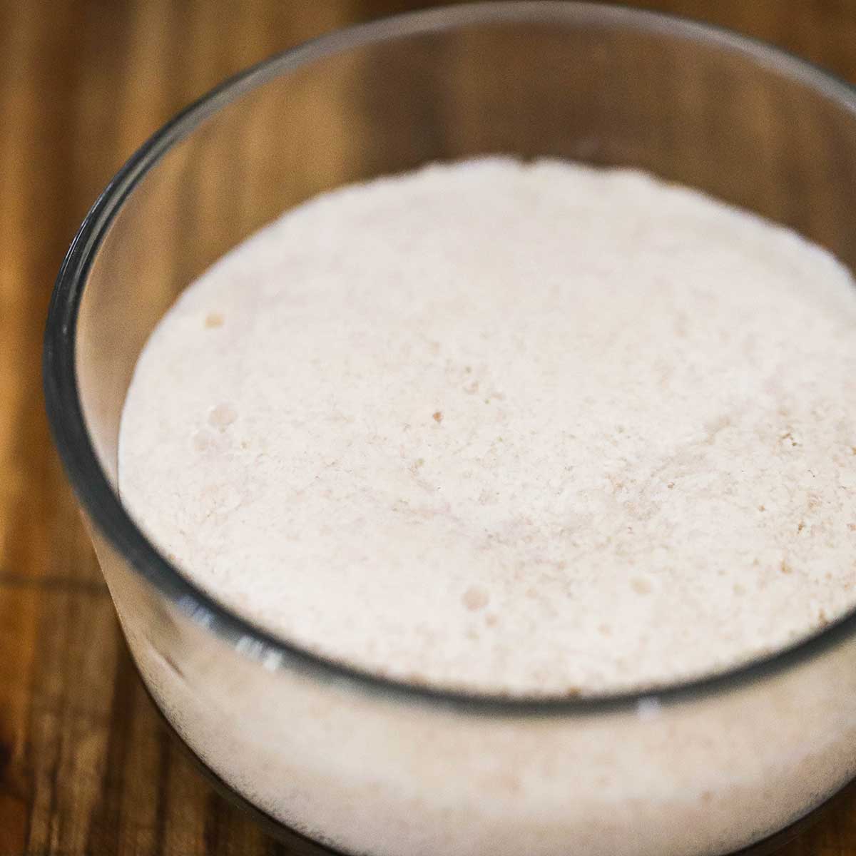 A small glass bowl filled with foamy yeast, sugar, and warm water on a cutting board. 