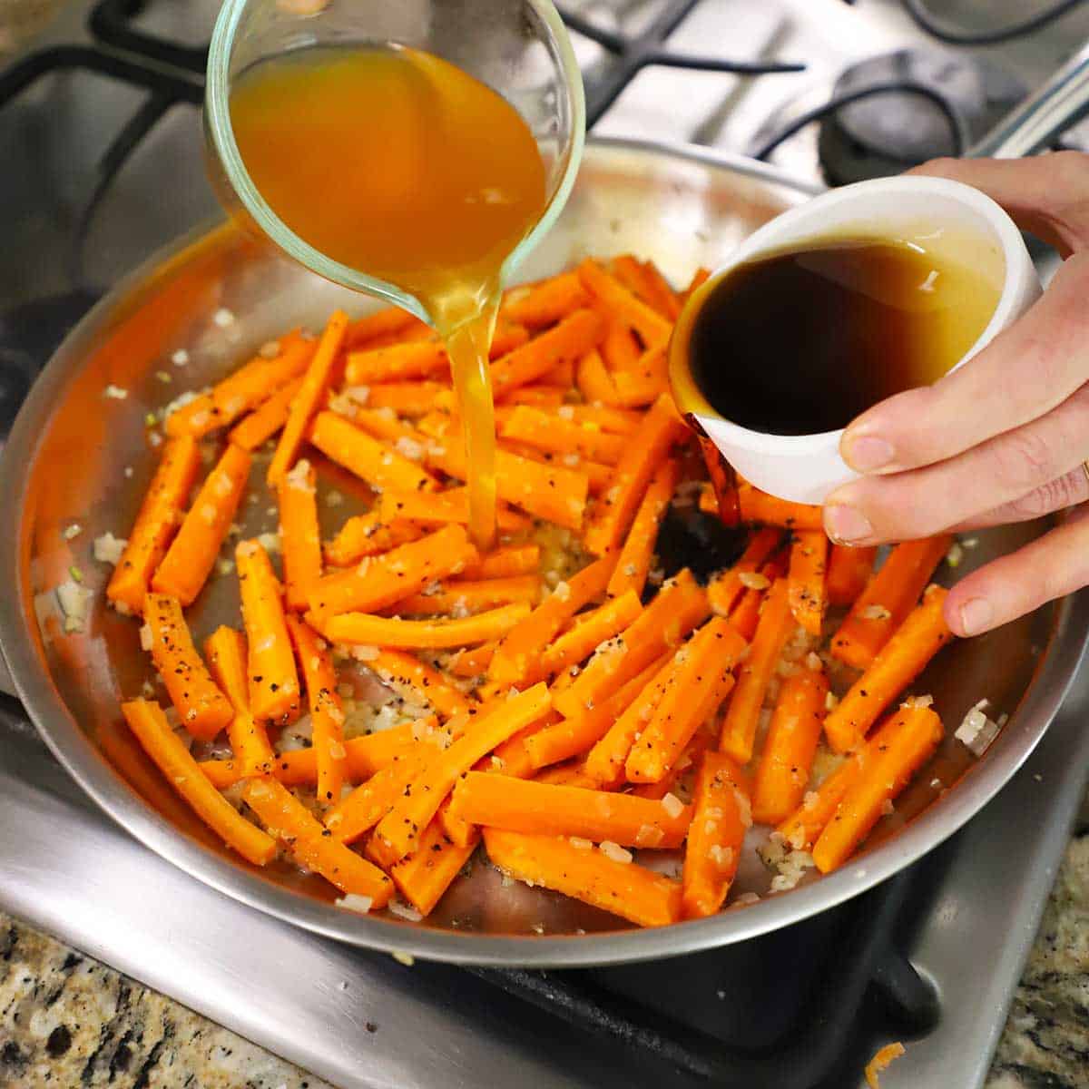 A person adding maple syrup from a small bowl and chicken broth from a measuring cup into a skillet filled with carrots strips.