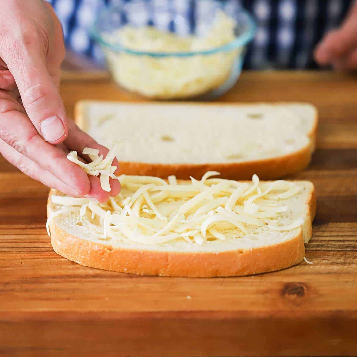A person sprinkling grated Gruyere cheese on to the top of a slice of sourdough bread that has a mayonnaise and Dijon spread on it. 