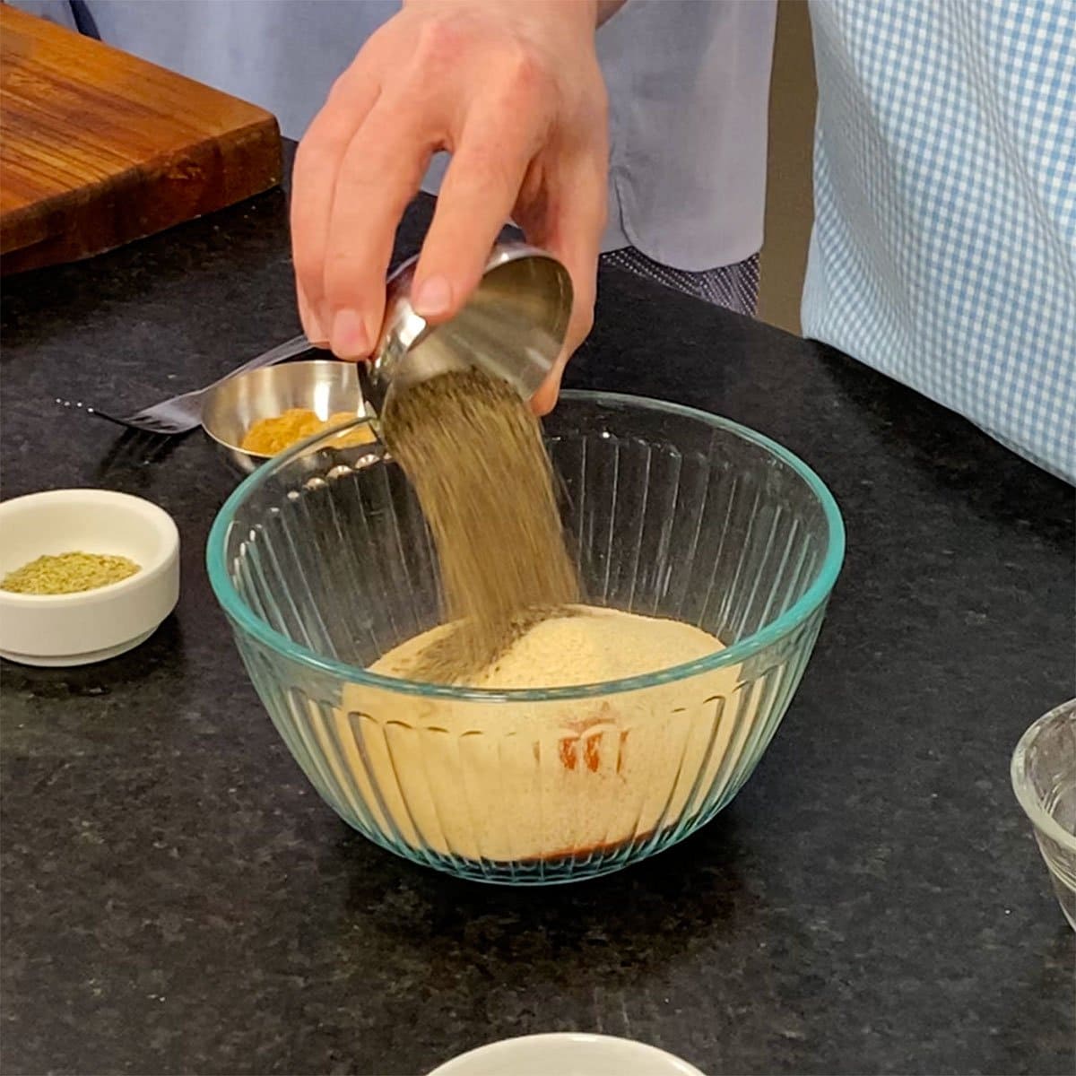 A person dumping ground black pepper from a small metal bowl into a glass bowl filled with spices including garlic powder, onion powder, paprika, and sugar.