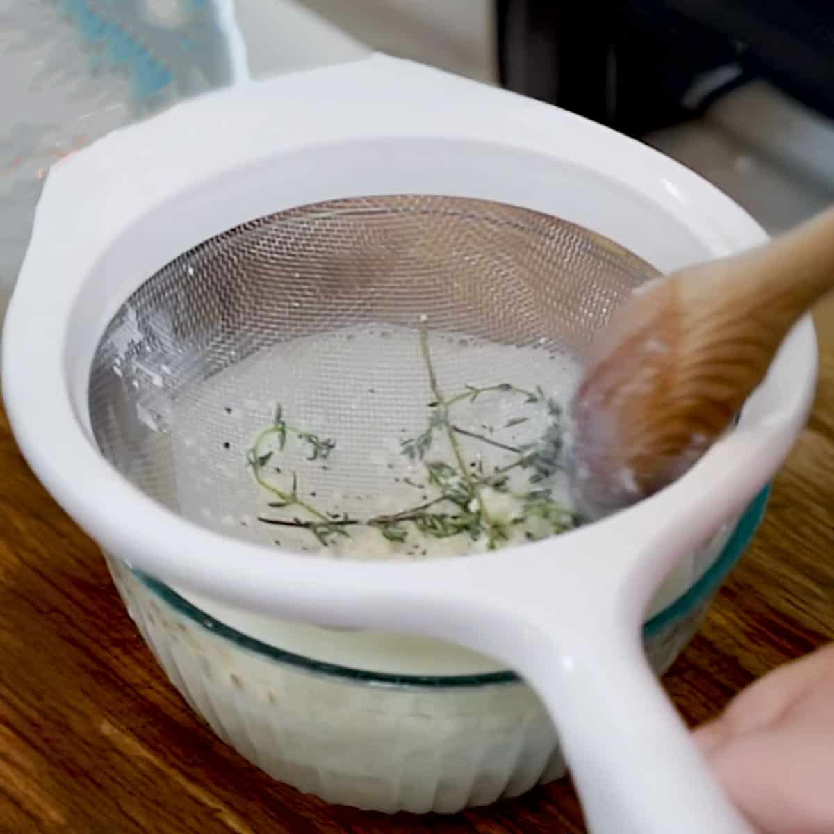 A person using a wooden spoon to press herbs in a strainer that is resting over a glass bowl filled of the cream mixture that had just been strained. 