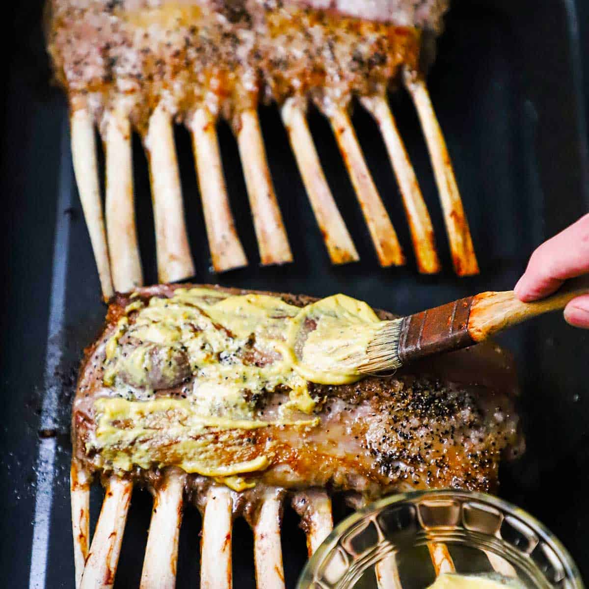 A person brushing Dijon mustard onto the top of a roasted rack of lamb that is in a roasting pan.